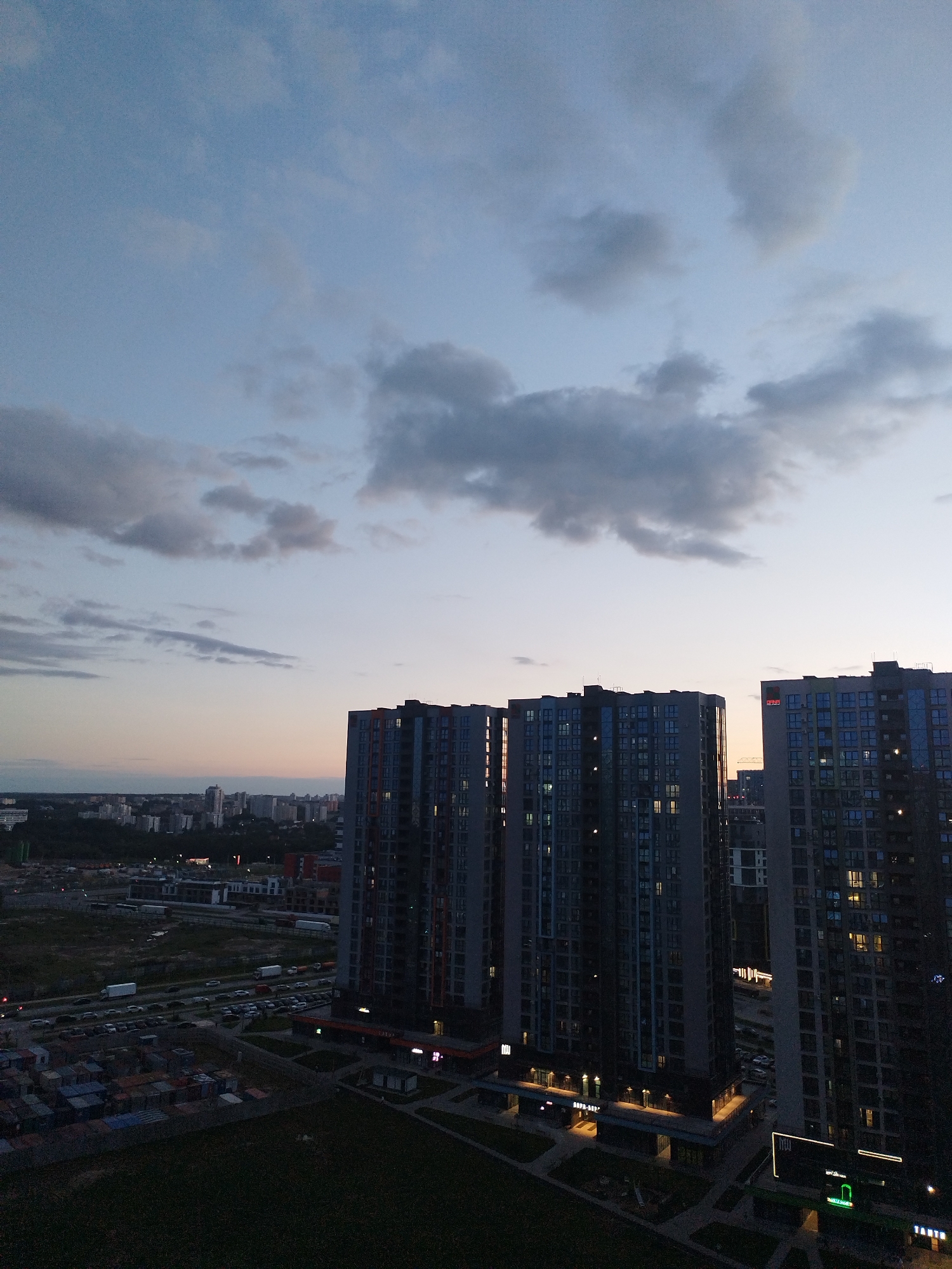 The image captures an aerial view of a city skyline at dusk. The sky is a gradient of soft blues and purples, with scattered grey clouds. Dominating the foreground are several tall, modern apartment buildings, characterized by their dark facades and numerous windows, many of which are illuminated, suggesting that people are home. The architecture is sleek and contemporary, with balconies and angular designs.
Below the apartment buildings, the ground level reveals a complex urban landscape. There's a busy road with a significant amount of traffic, indicated by the lines of cars and trucks. Adjacent to the road are parking lots filled with vehicles, and further down, a collection of shipping containers is stacked, suggesting commercial or industrial activity. Beyond the immediate vicinity of the buildings, the cityscape extends into the distance, with a dense collection of other buildings of varying heights, hinting at a large, populated metropolitan area. Trees and green spaces are visible in some parts of the landscape, offering a contrast to the concrete structures.
The time of day is clearly evening, with the fading sunlight casting a soft glow on the horizon and the artificial lights of the buildings beginning to stand out. The weather appears to be clear, with the clouds being dispersed and not indicating any immediate precipitation. The overall mood is one of a transition from day to night in an active urban environment.
Visible text includes signage on the ground-level storefronts of the apartment buildings. One sign reads "SOFA-BAR" in illuminated lettering. Another sign appears to say "TASTO". There is also a neon green sign with a light bulb graphic. The text on the buildings themselves is minimal and generally not legible from this distance.