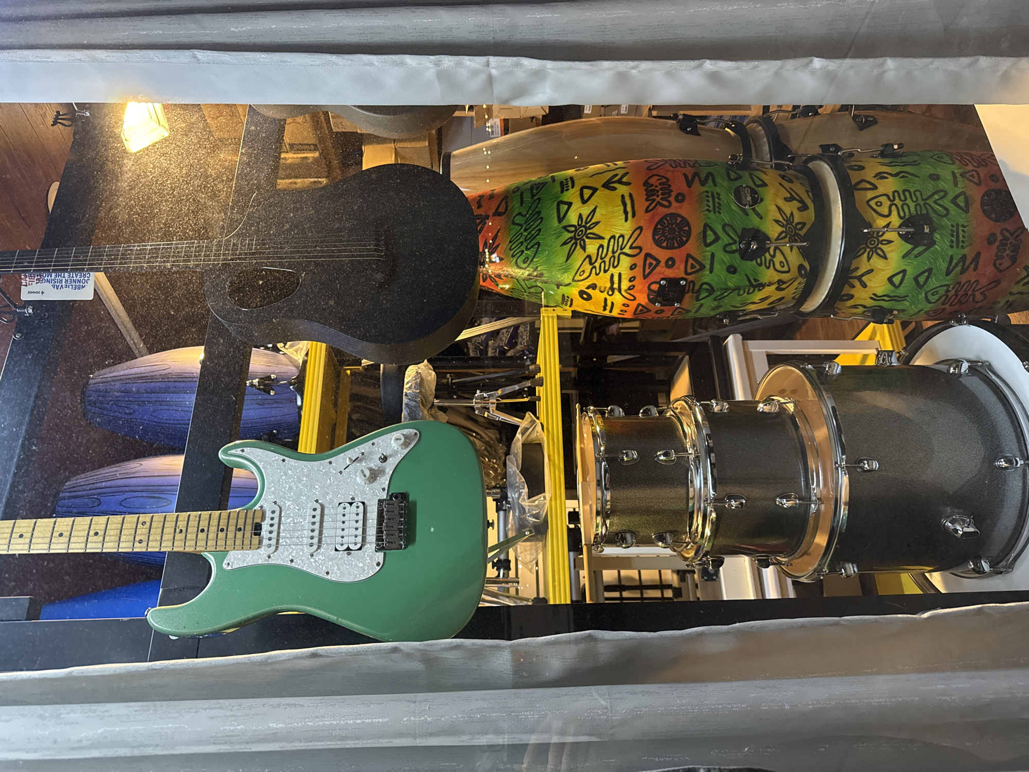 The image displays a collection of musical instruments, including guitars and drums, arranged in what appears to be a display or storage area. A black electric guitar with a distinct soundhole and a green Fender Stratocaster are prominently featured. To the right of the guitars are several drums, including two conga drums decorated with a vibrant, tribal-inspired pattern in shades of red, yellow, and green, featuring abstract designs and fish-like motifs. Further to the right, a set of metallic grey drums, possibly a drum kit, is visible. In the background, out-of-focus elements suggest a cluttered environment with wooden flooring and lighting fixtures. One of the guitars has a sticker on its body with the text "BELL-VAN DONNER RISING CREATE THE N..." visible. The lighting in the scene is warm and slightly dappled, suggesting an indoor setting, possibly a shop or a music studio. The overall impression is one of a display of musical equipment.