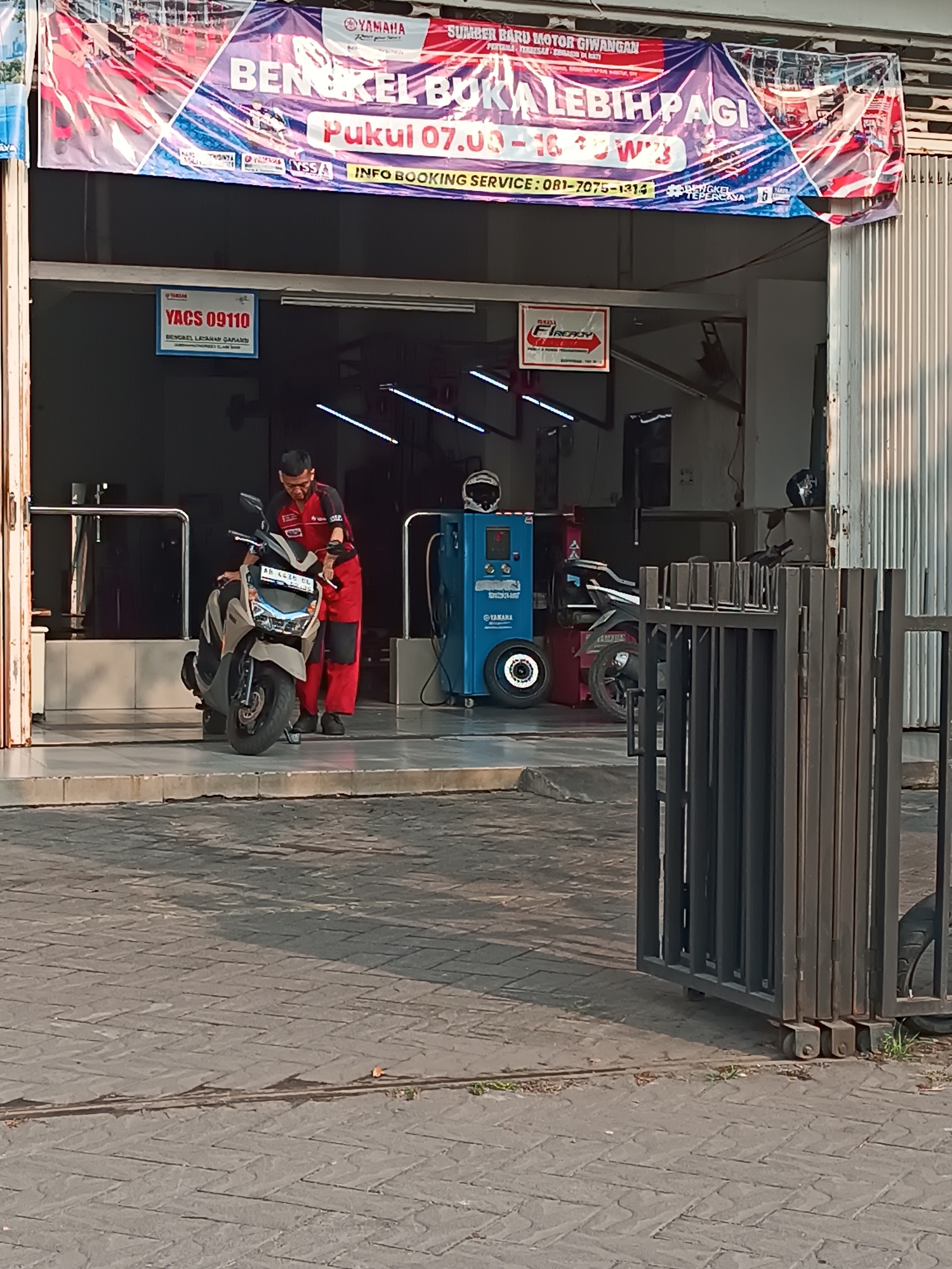 The image shows the exterior of a Yamaha motorcycle repair shop in Banguntapan, Indonesia. A man in a red and black uniform is standing next to a beige scooter, appearing to be inspecting it. The shop has a large banner hanging above the entrance with the words "BENGKEL BUKA LEBIH PAGI" (Workshop Opens Earlier) and "INFO BOOKING SERVICE: 081-7075-1314". There are also signs for "YACS 09110" and "FIREADY". In the background, a blue service machine with a tire attached is visible. The ground outside the shop is paved with interlocking bricks, and a metal gate stands in the foreground. The scene appears to be captured during daylight hours.