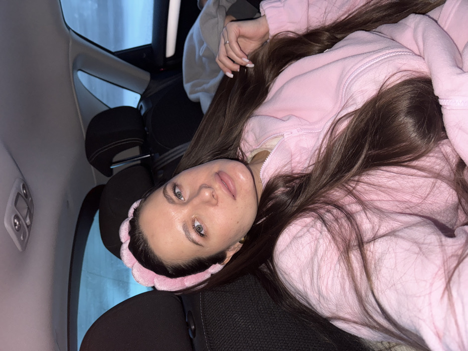 This image depicts a single female subject situated in the back seat of a vehicle in Lavrovo, Russia. The subject, a woman with long, wavy dark brown hair, is wearing a light pink, textured headband and a light pink zippered fleece jacket over a lighter garment. Her light-colored eyes are directed towards the camera, and her lips appear lightly enhanced. Her left hand, adorned with a silver-colored ring on the ring finger and manicured white fingernails, rests near her hair on the right side of the frame. The vehicle's interior features dark upholstery on the headrests and seatbacks, with a light-colored roof liner and an overhead light fixture visible. Through the car windows, a blurred, pale blue and white outdoor environment is discernible, consistent with snow or an overcast winter sky. The overall lighting suggests low light conditions, possibly dusk or night.