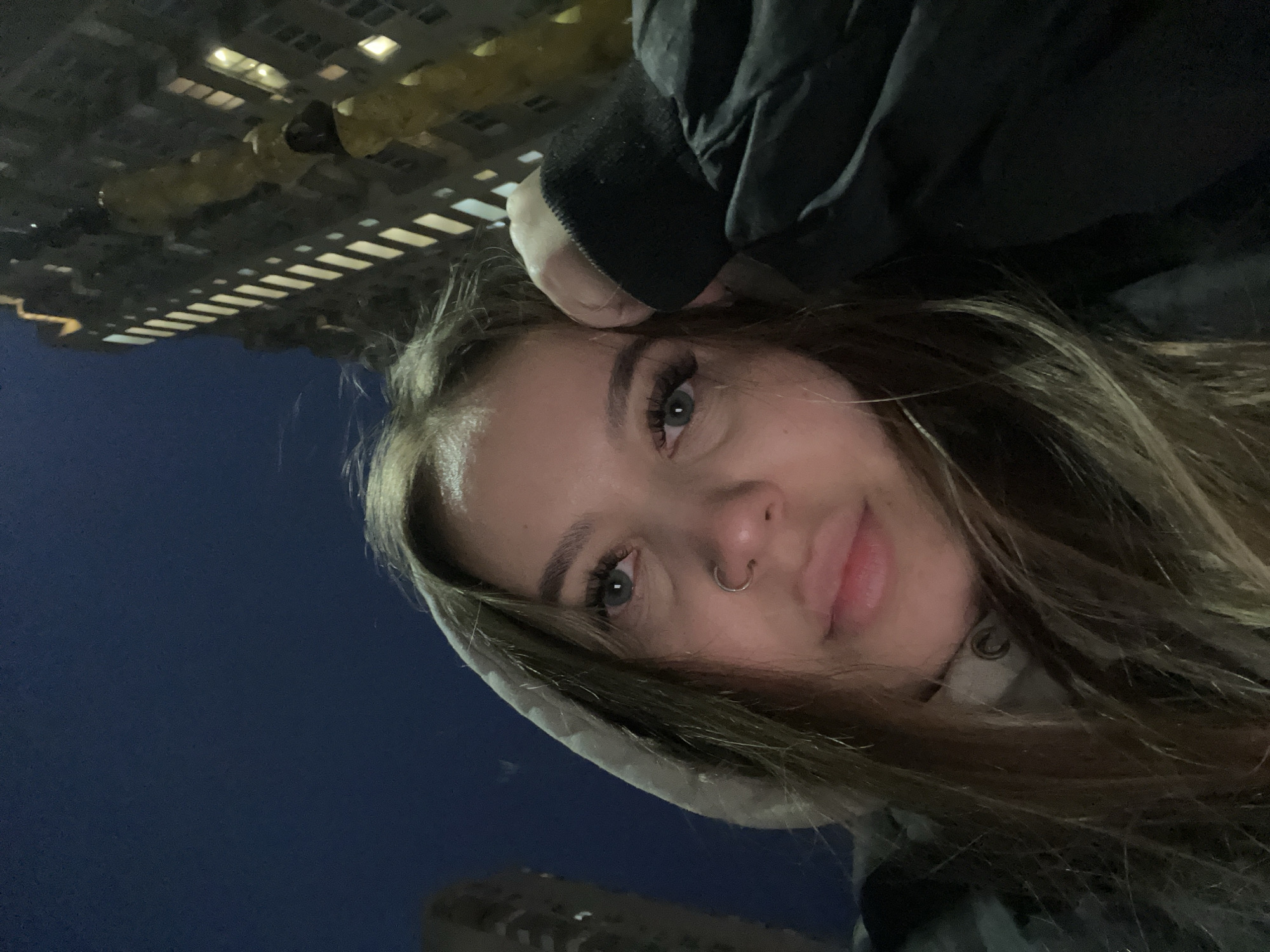 A close-up selfie of a young woman with light brown hair, wearing a dark jacket with a hood, is oriented vertically. She has fair skin, prominent eyebrows, long eyelashes, and a nose ring. Her left hand is visible near her forehead. The background is dark blue, suggesting night. Reflected lights from buildings are visible at the top and bottom of the image, indicating an urban setting. Based on the architecture and lighting patterns, this could be Krasnodar, Russia.