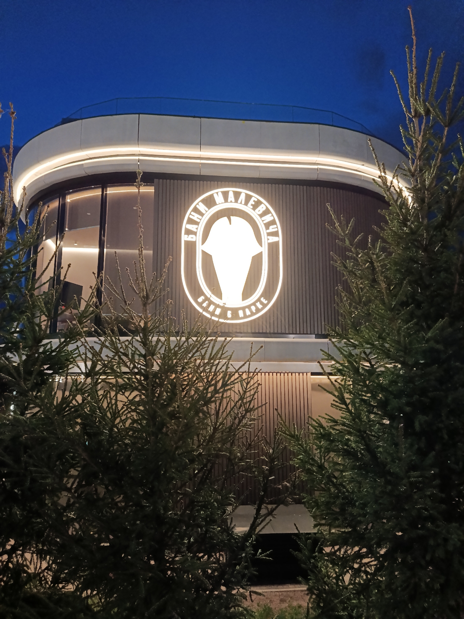 Captured at night in Razdory, Russia, the image features a modern two-story building with a rounded facade. The building's upper section is illuminated by a perimeter light. A large, oval-shaped sign with neon lighting prominently displays the words "Бани Малевича" (Bani Malevicha) in Cyrillic script, framing a stylized icon resembling a bathhouse-related object. The building has vertically aligned exterior panels. In the foreground, evergreen trees partially obscure the view of the building, adding depth and a natural element to the composition. The sky is a deep blue gradient, suggesting twilight or early evening.