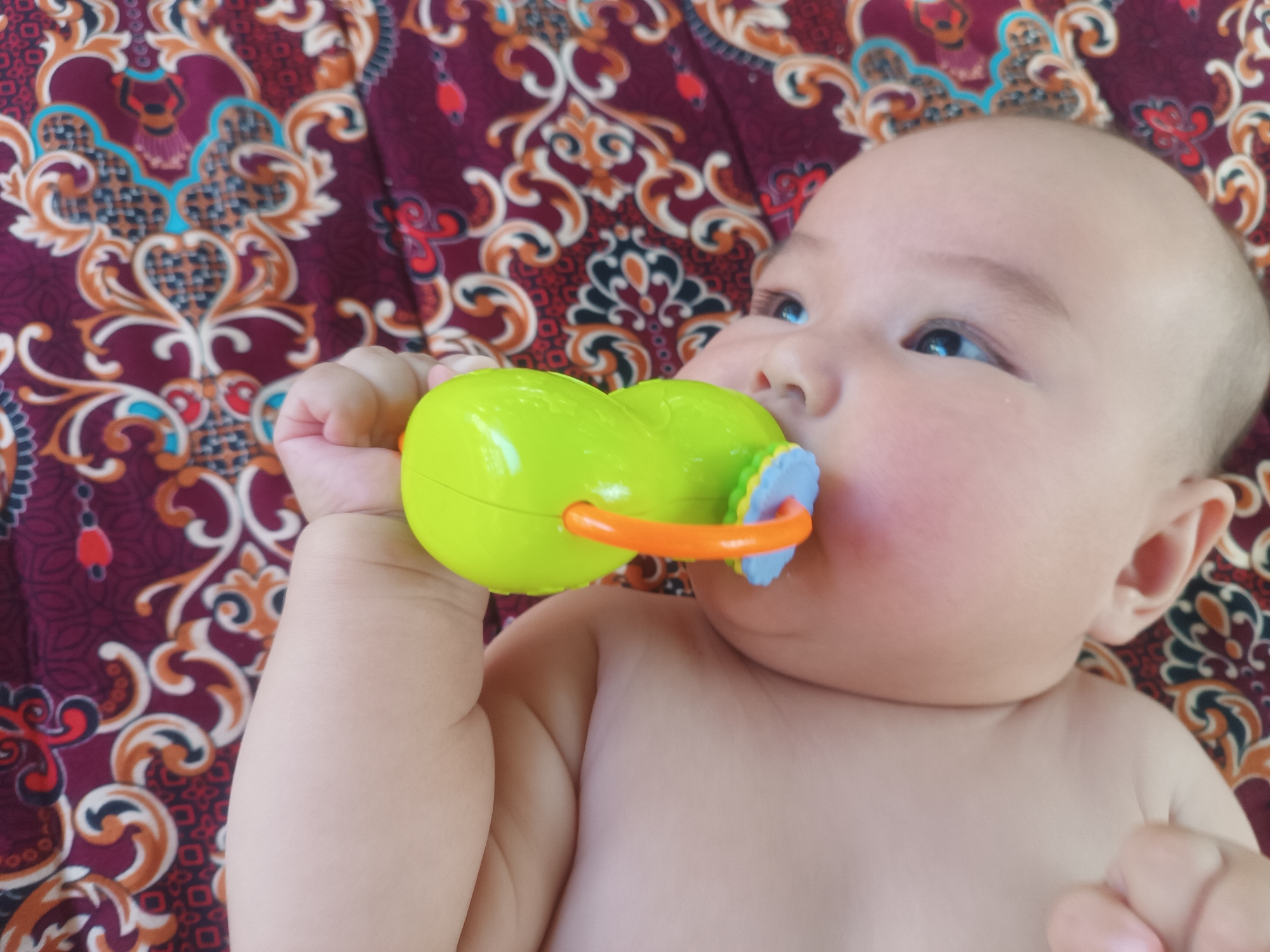 A close-up, overhead shot shows a bald baby lying on a patterned fabric, possibly a blanket. The baby is looking to the right, with its eyes open. The baby is holding a bright green toy in its right hand, which is partially in its mouth. The toy is shaped like a peanut and has an orange ring and a blue and white textured piece. The baby's skin appears fair, and its cheeks are slightly flushed. The fabric beneath the baby features a detailed pattern in shades of maroon, orange, cream, and blue, with swirling floral and geometric motifs. The background is entirely filled with this fabric. The setting is domestic. The image does not provide any specific geographical markers that would indicate the location in Myrza-Ake, Kyrgyzstan.