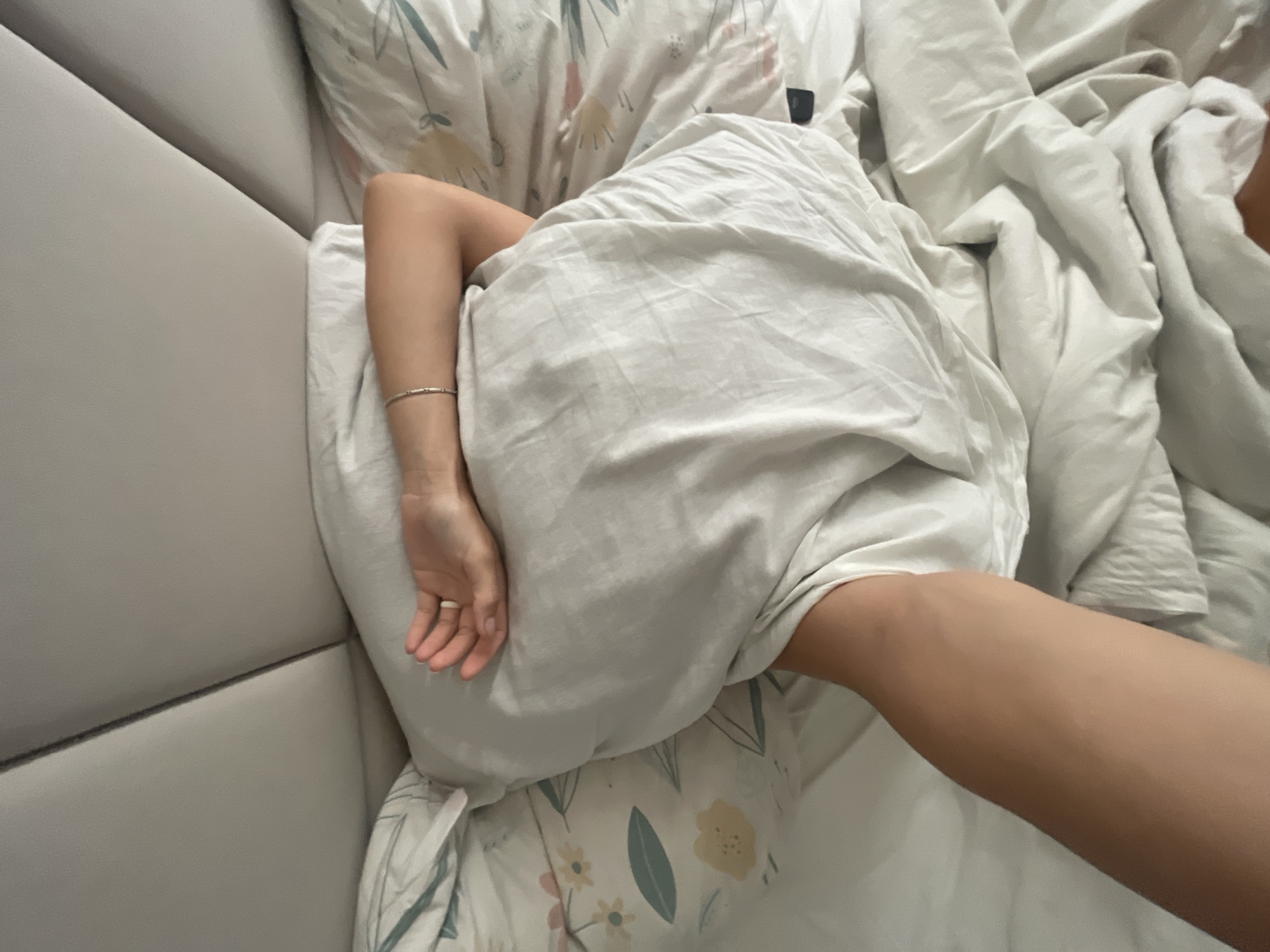 The image displays an overhead perspective of a person resting in bed, likely indoors in Baranovka, Russia. The scene captures a moment of repose, with the individual largely covered by bedding.
The central subject is a person, whose left arm and part of a leg are visible. The arm is bent, resting on a pillow, with a hand visible featuring a simple ring on one finger and a thin, silver-colored bracelet on the wrist. A portion of the person's leg or thigh, with fair skin, emerges from under a white sheet on the right side of the frame.
The bed is adorned with light-colored bedding. A plain, light grey or white sheet covers the person's body, exhibiting natural folds and wrinkles. Underneath and around the person's arm and head are pillows covered with patterned pillowcases, featuring a delicate design of green leaves and pale yellow or white flowers. To the far right, additional crumpled white bedding, possibly a duvet or another sheet, is visible.
To the left of the frame, a segment of a light grey upholstered headboard is prominent. It has a clean, modern aesthetic with a distinct horizontal seam running across it. The overall setting suggests a cozy and comfortable bedroom environment. No specific time of day is explicitly indicated, but the scene implies a period of rest or sleep. There is no visible text within the image.