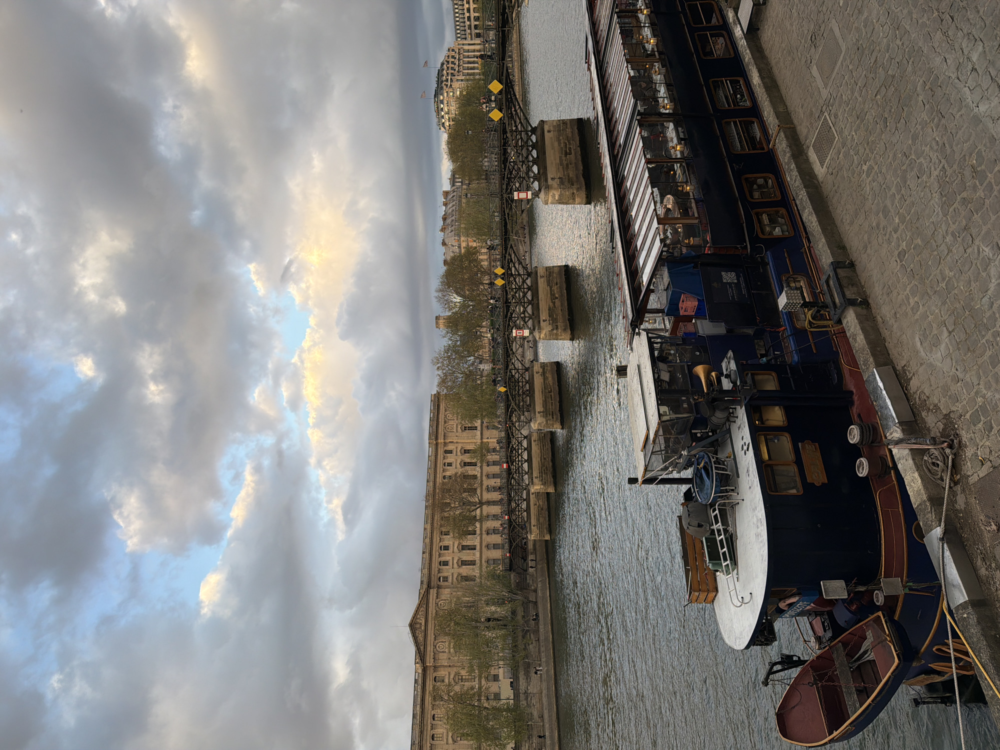 This picture captures a view of Paris, France. A river flows through the scene, and a pedestrian bridge crosses it. A large barge-like boat is moored on the right side of the river. The architecture of buildings on the left river bank is distinctly Parisian. The sky is filled with dramatic clouds. The image is shot at a slight angle.