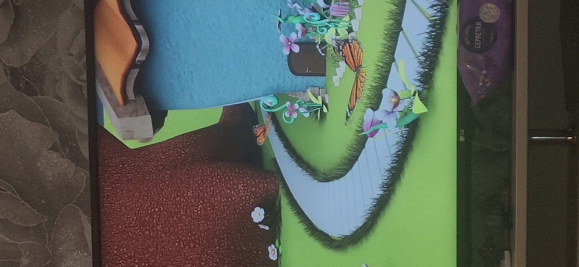 The image is a close-up of a television screen displaying a cartoonish, animated scene. The scene features a bright green grassy landscape with a winding, light blue pathway made of square stones. To the left, a large, textured, reddish-brown object resembling a tree trunk dominates the background. To the right, a small, blue house with an orange roof is visible, with a dark brown door. In front of the house and along the pathway, there are various colorful, cartoon-style flowers in shades of pink and purple. Two orange butterflies with black markings are present in the scene, one on the pathway and another near the base of the tree trunk. The bottom of the television screen shows the LG logo and a portion of a purple package with Russian text that appears to read "СЕРБЕТКИ" (servetki), meaning "wipes" or "napkins." The setting is an outdoor, whimsical environment. The image does not contain any information about the city of Bila Tserkva, Ukraine.
