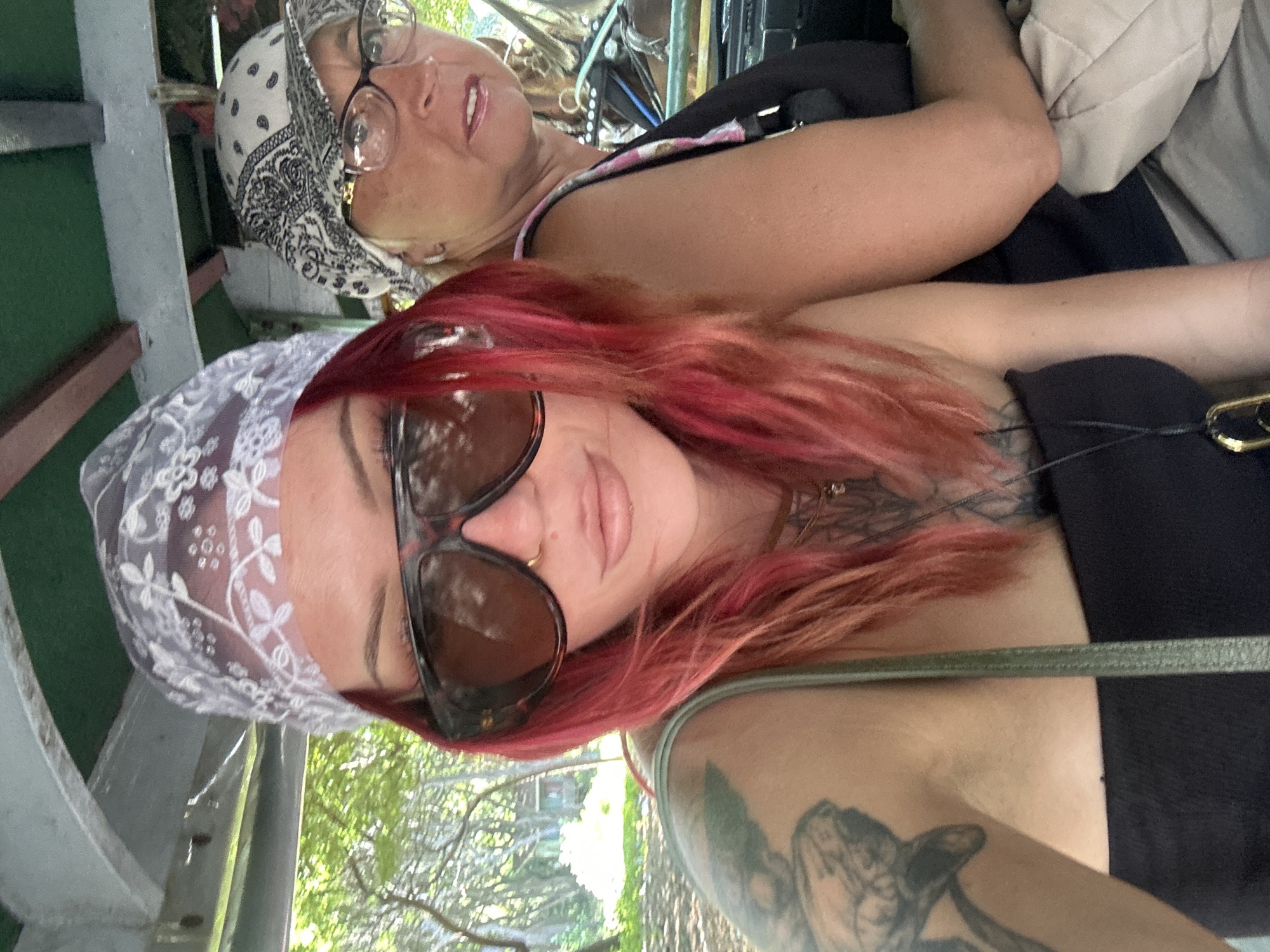 Two women are sitting inside a vehicle. The woman in the foreground has red hair and is wearing a lace headscarf and sunglasses. She has tattoos on her neck and arm. The woman behind her is wearing a bandana and glasses. The location is the city of Unknown, Indonesia. Outside the vehicle is greenery.