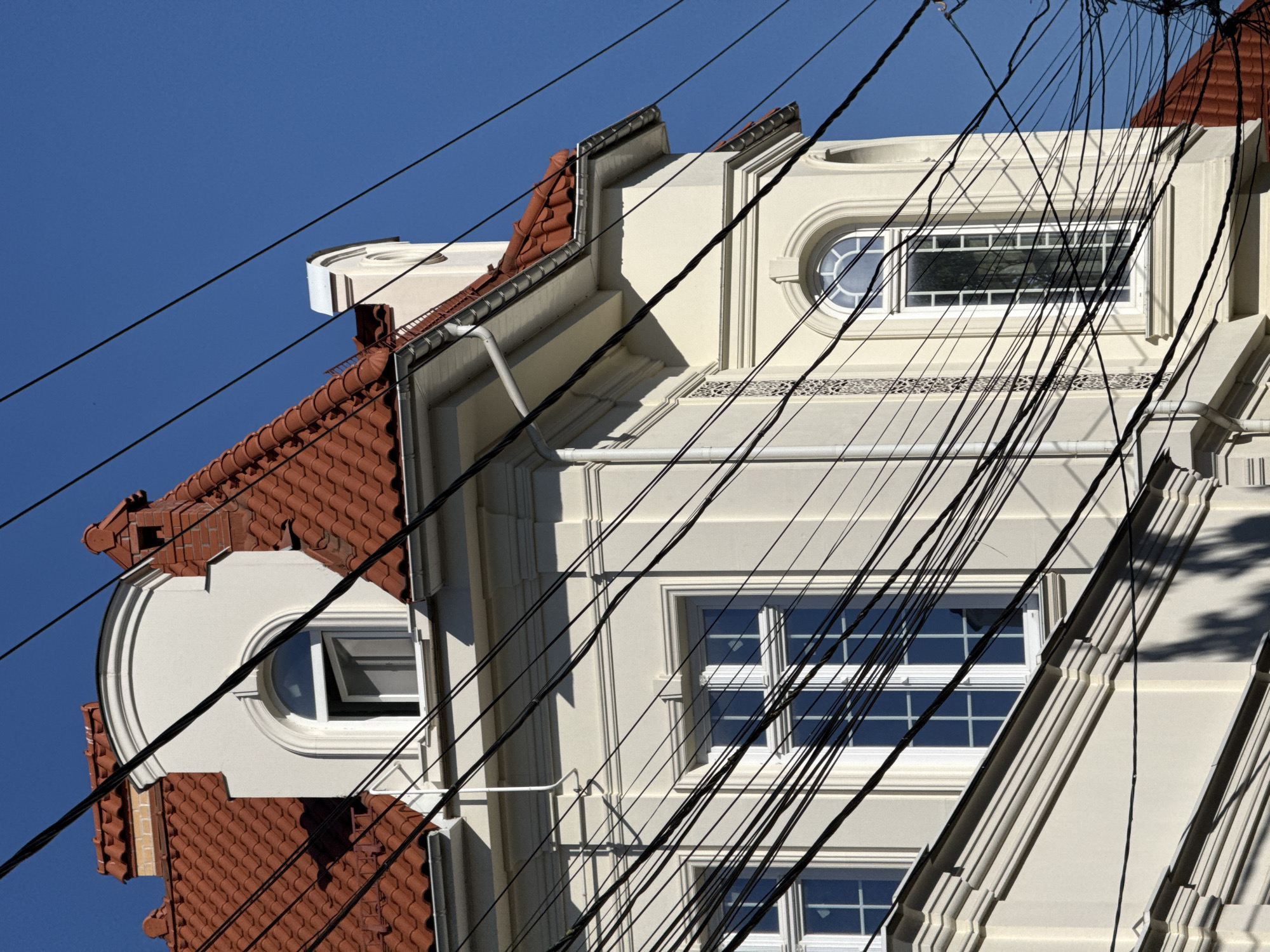 The photograph captures a close-up, oblique view of the upper floors and roofline of a building in Odesa, Ukraine. The building's facade is cream-colored with ornate white trim around its windows. It features several white-framed windows, including a prominent arched window with multiple panes and a dormer window with an open sash. The roof is covered in reddish-orange tiles and has a stepped gable design. A complex network of black electrical wires crisscrosses the foreground, obscuring parts of the building and the clear blue sky. The lighting suggests it is daytime, with shadows cast by the wires and architectural elements. No people are visible in the frame.