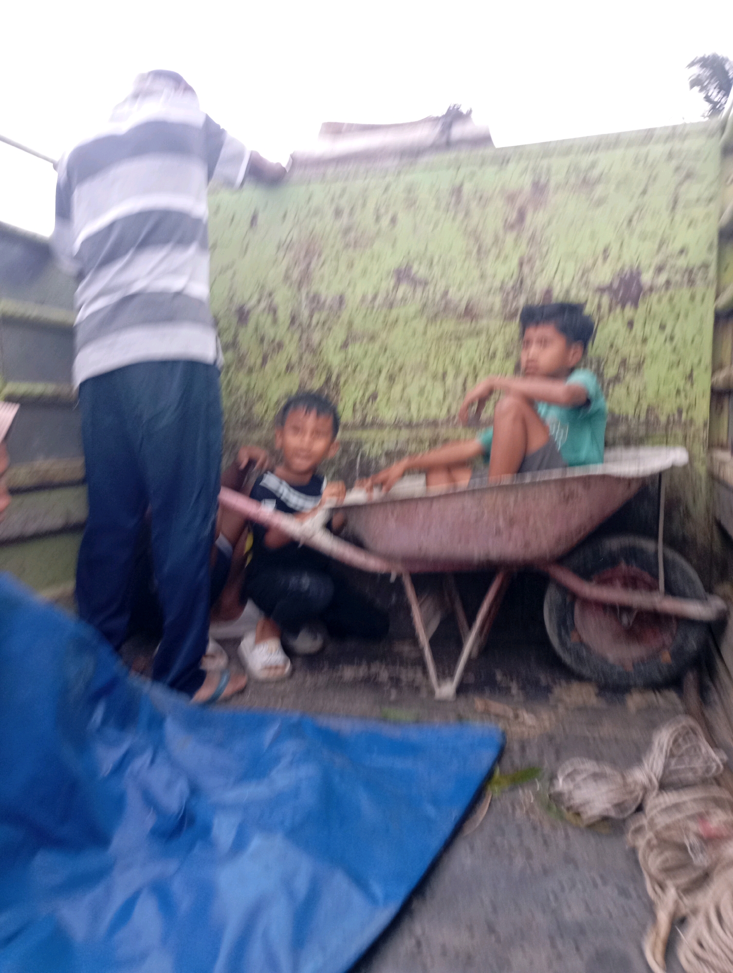 The image, taken in Menadi, Indonesia, shows a group of people in the back of a truck. In the foreground, a blue tarp is spread out. To the left, a person wearing a striped t-shirt and blue pants stands facing away from the camera, holding onto the side of the truck. In the center of the truck bed, two young boys are visible. One boy, wearing a black and white striped shirt and dark pants, is sitting in a wheelbarrow. The other boy, wearing a teal t-shirt and dark shorts, is also sitting in the wheelbarrow, leaning against the side of the truck. The truck bed is green and appears to be made of metal. To the right of the wheelbarrow, a rope is coiled on the floor of the truck. The background is out of focus but appears to be an outdoor environment with some greenery visible at the top.