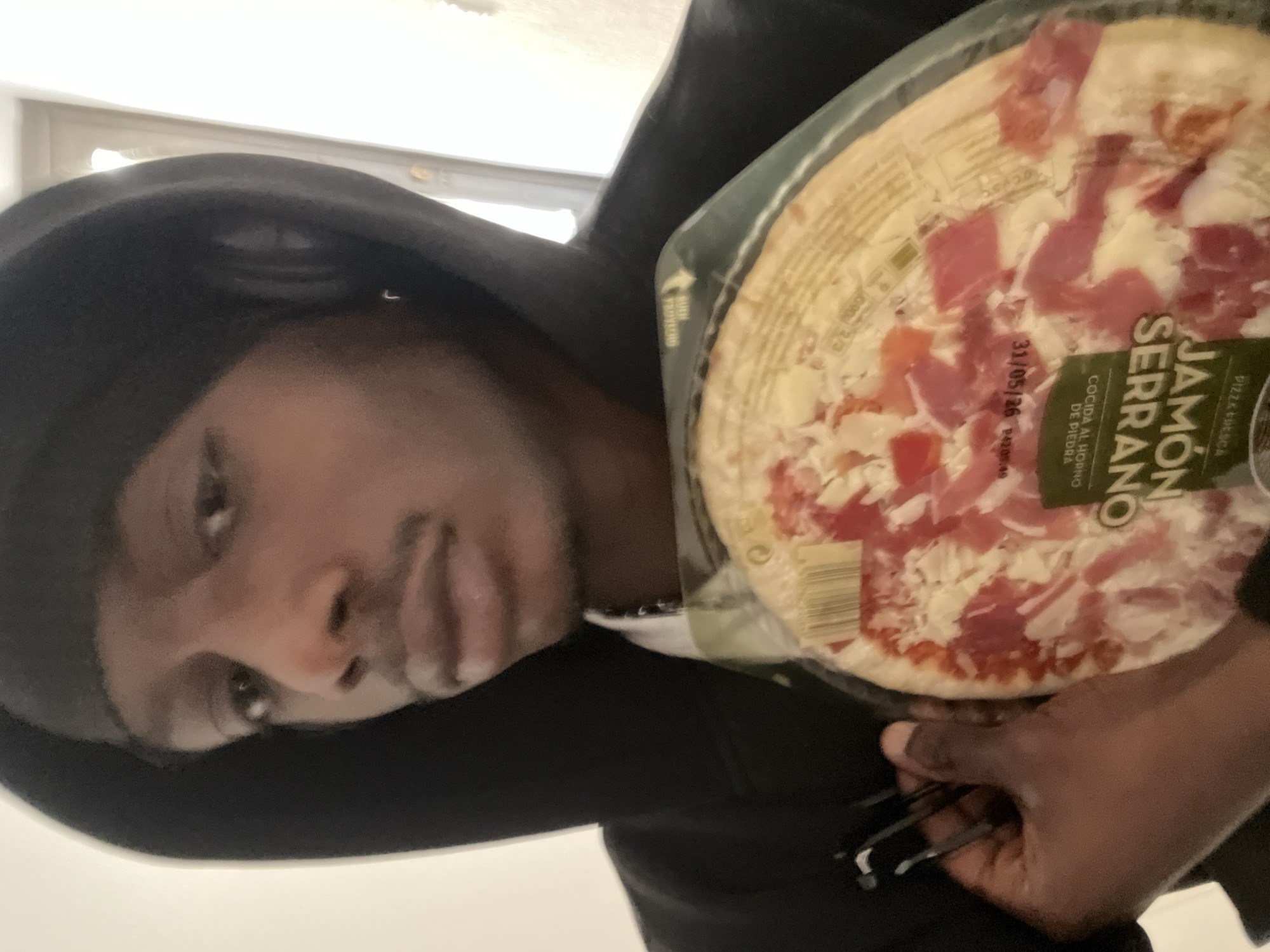 A person wearing a black hoodie holds a packaged pizza in front of them. The pizza's packaging is partially labeled "Jamón Serrano" and displays an expiration date of "31/05/26". The location in Barcelona, Spain is unknown.