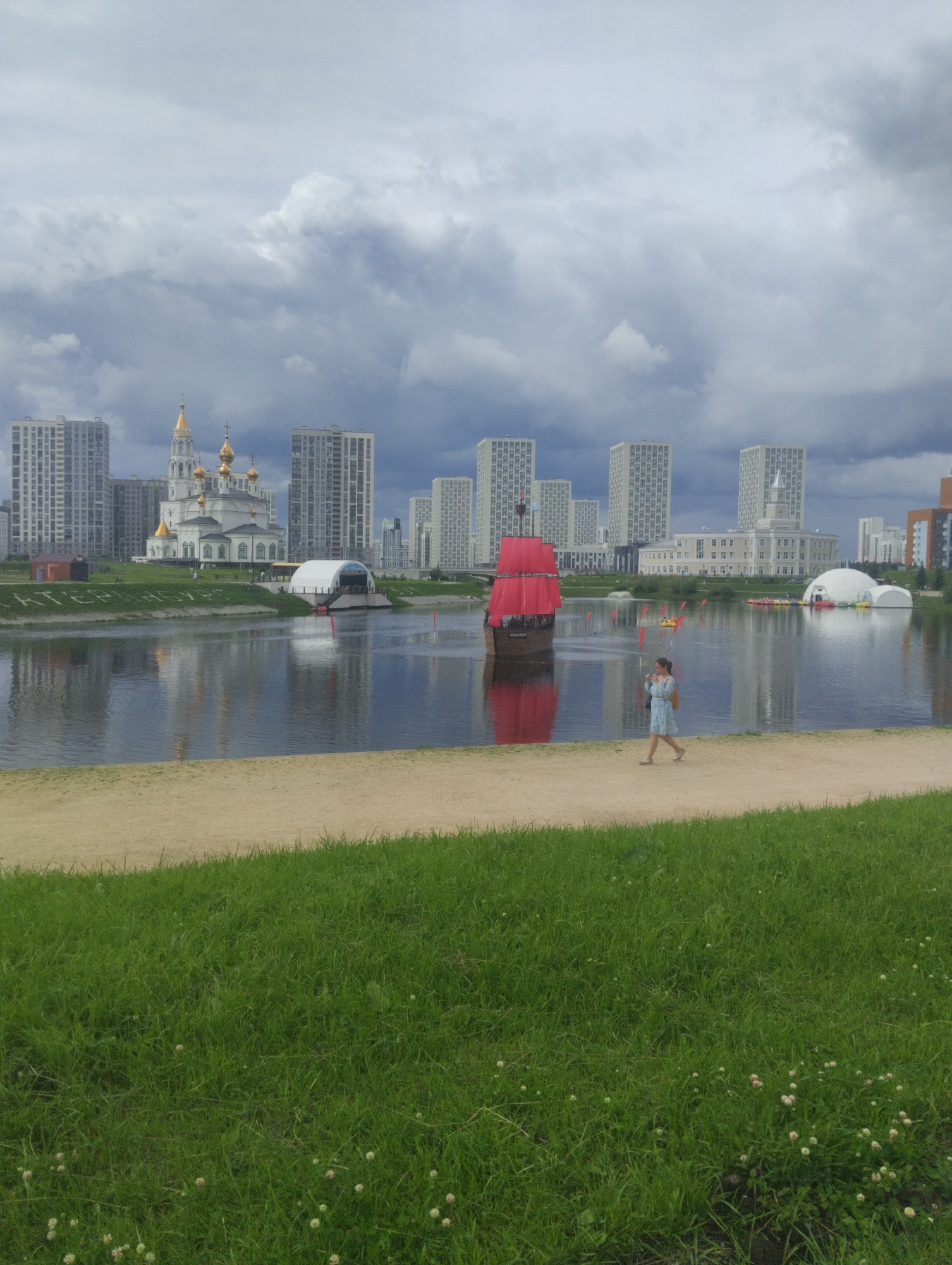 This image captures an outdoor scene on an overcast day, likely in a park or waterfront area of a city. In the foreground, a grassy slope descends to a sandy path. Beyond the path, a body of water, possibly a river or lake, reflects the sky and the surrounding structures. On the water, a replica of a pirate ship with red sails is prominently featured. A woman in a light blue dress is walking along the sandy path, holding a white umbrella and a bag. In the middle ground, across the water, a white domed building is visible, as well as several modern, multi-story apartment buildings and a white building with a dome and columns. The sky is filled with dramatic, dark clouds.