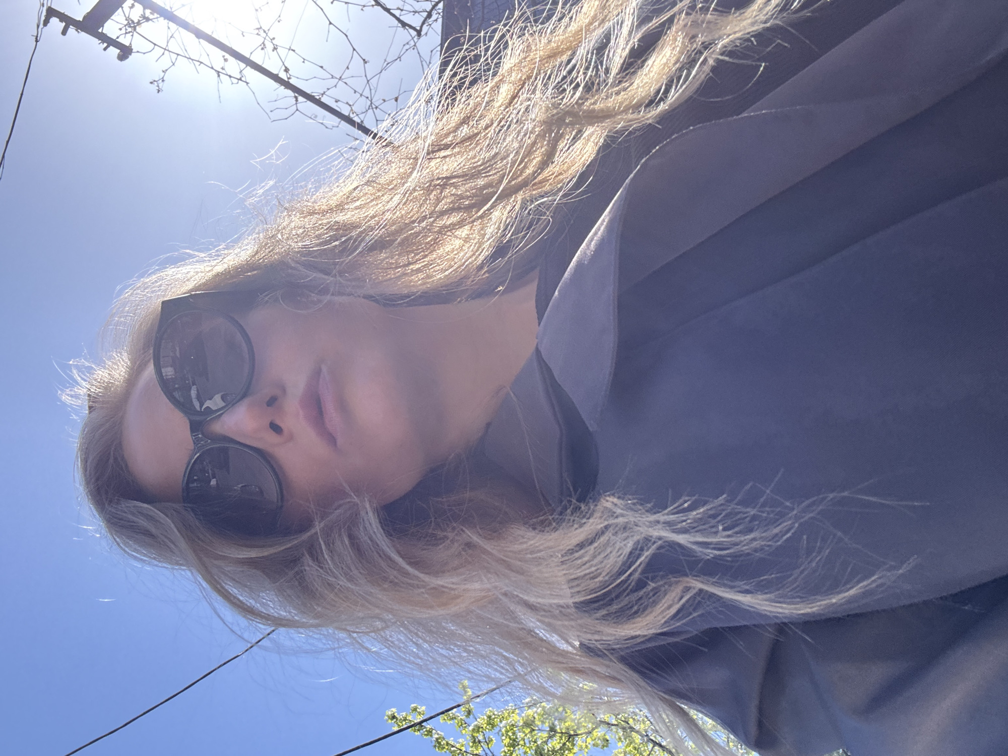 The image displays a low-angle outdoor portrait of a person, likely a woman, taken on a bright, sunny day. The subject is seen from below, with her face partially obscured by large, round, dark sunglasses. Her long, wavy, blonde or light brown hair cascades around her shoulders, catching the intense sunlight which creates a bright halo effect around it. She is wearing a dark blue or gray garment, possibly a jacket or shirt, visible on the right side of the frame. Her lips are slightly parted.
The background consists primarily of a clear, bright blue sky. A strong glare from the sun is visible in the upper left corner. Silhouetted against the sky are numerous bare tree branches, suggesting late winter or early spring. A thin black wire or cable stretches horizontally across the middle of the sky. In the lower right corner, a small patch of green foliage from a tree can be seen.
The photograph appears to be a selfie, capturing the subject looking towards the camera from a downward angle. The lighting is very bright, casting some areas of the subject's face into shadow due to the backlighting. There is no visible text in the image.