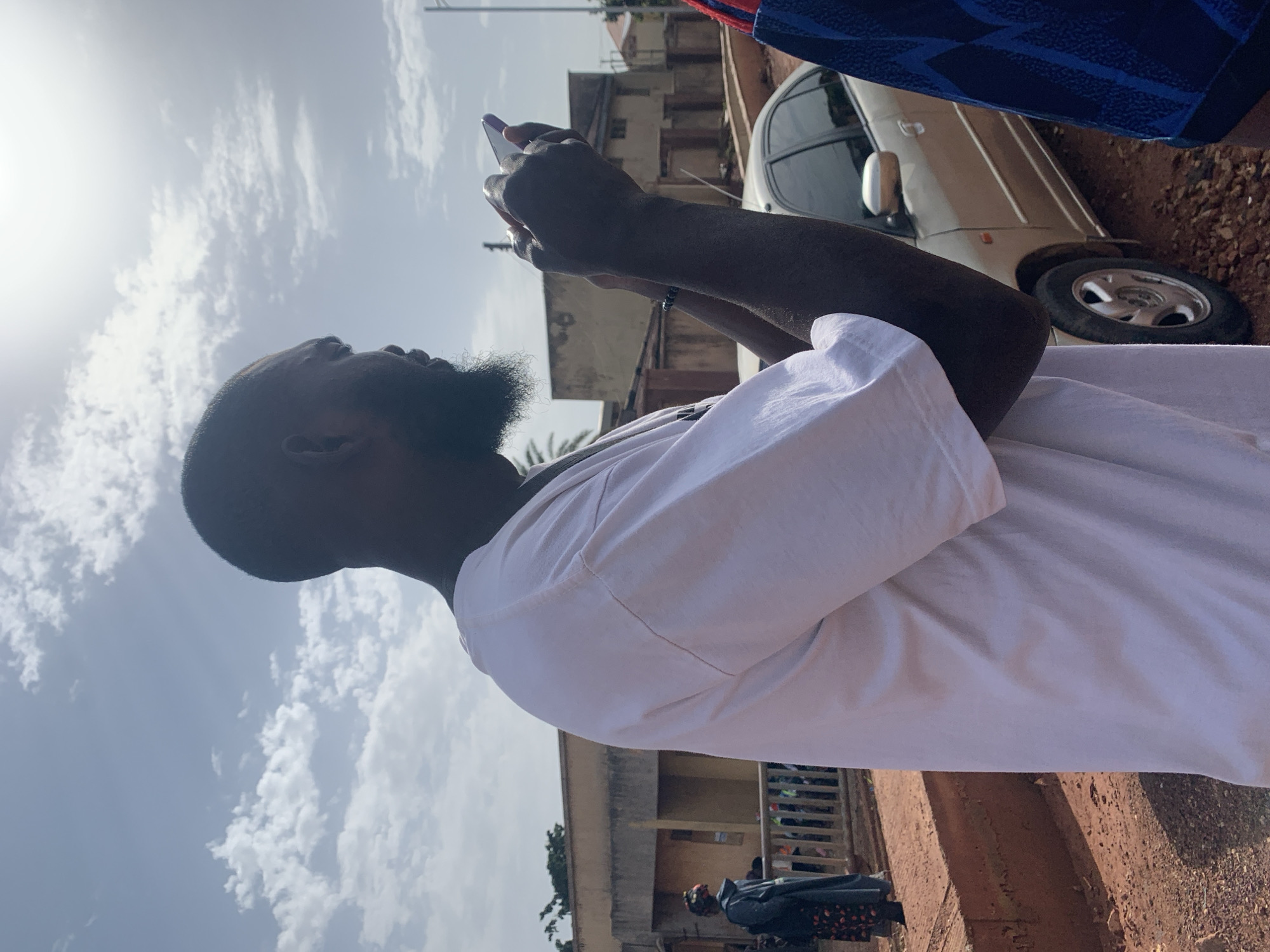 The image shows a man with a beard wearing a white t-shirt, standing outdoors and looking at his phone. He is in a crouched position, holding the phone with both hands. In the background, there are buildings, a fence, a car, and other people. The ground is covered in dirt and gravel, and the sky is visible with clouds and sunlight. The scene appears to be a street or an open area in a residential or commercial district. The location is identified as Gura Top, Nigeria.