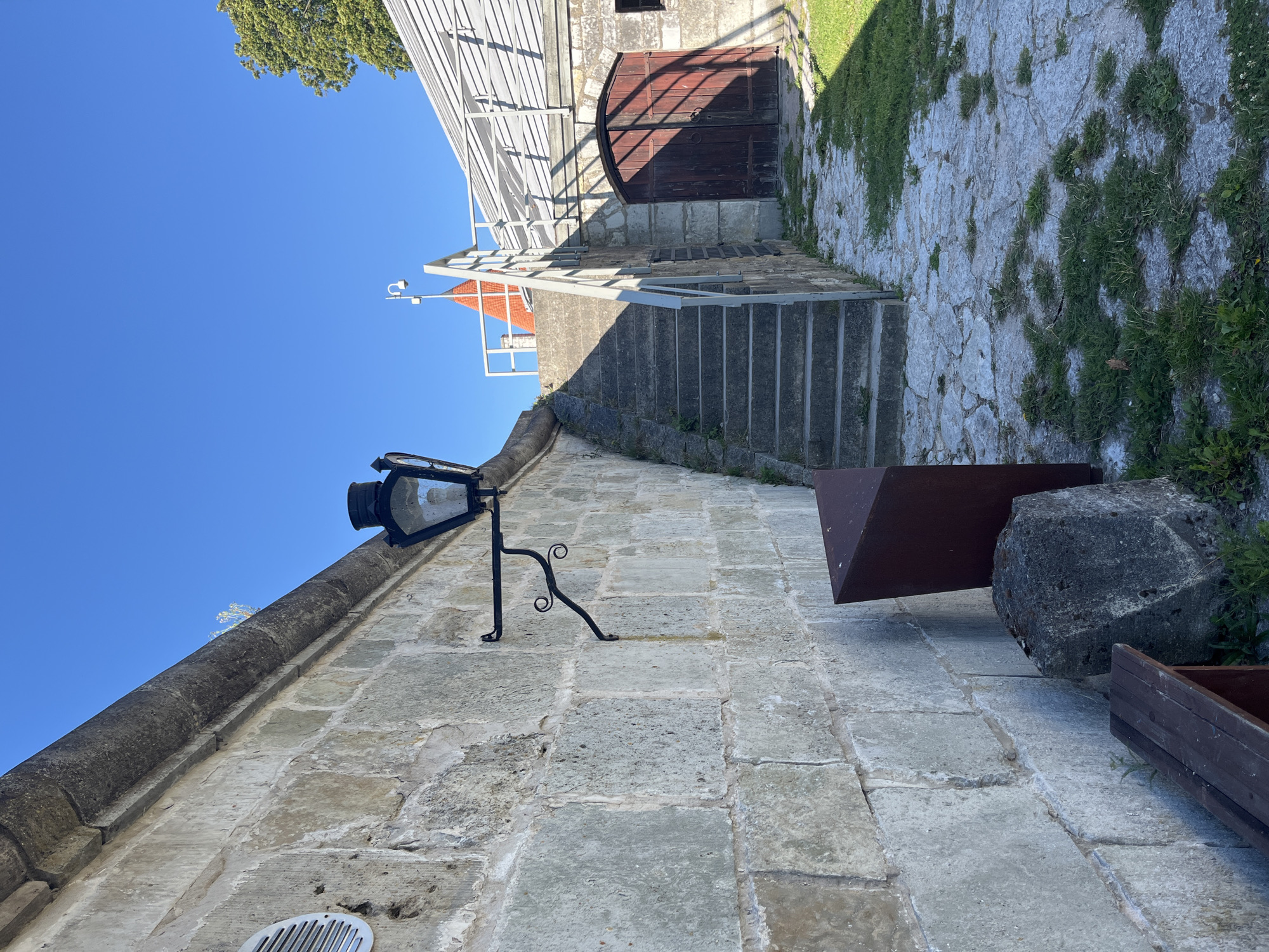 This is a photograph taken outdoors in the city of Kuressaare, Estonia. The scene is dominated by a weathered stone wall on the left side of the frame. Attached to this wall is an ornate black metal lamppost with a glass enclosure. To the right of the wall, a set of concrete stairs ascends towards a building with a sloped, white roof. Adjacent to the stairs is a large, weathered wooden double door. The ground is a mix of flagstone paving with patches of green grass growing through the cracks. In the foreground, a dark wooden bench is visible. The sky is a clear, bright blue.