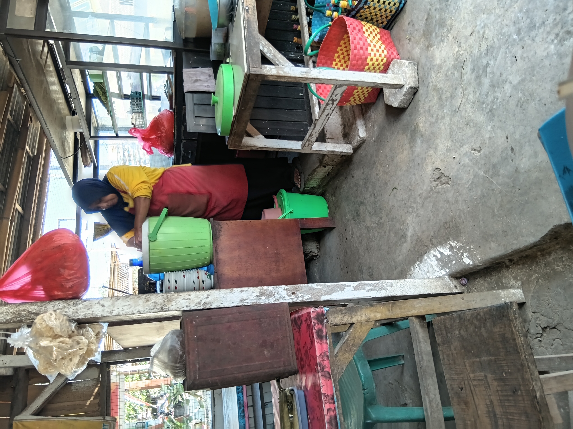 The image depicts an outdoor food stall in Kasiyan Timur, Indonesia, likely in a residential or market area, with a focus on food preparation and sale. A woman, wearing a yellow long-sleeved shirt, a red apron, and a dark hijab, is actively engaged in food preparation. She is standing behind a wooden counter, and her hands are near a large green plastic cooler.
The stall appears to be a simple, open-air structure with a corrugated metal roof supported by wooden beams. Various items associated with food service are visible. To the left of the woman, a large red plastic bag is hanging, and above it, a cluster of pale, dried food items, possibly crackers or snacks, are also suspended in plastic wrapping. A dark-colored, rectangular metal container sits on a red-patterned table to the left.
The floor of the stall is concrete, showing signs of wear and some white paint splatters. Several plastic buckets and containers are scattered around the area, including the prominent green cooler and a smaller green bucket near the woman's feet. In the background, shelves are visible, stocked with various items, some of which appear to be food products or kitchen supplies. Woven baskets, one red and yellow and another blue, are placed beneath a wooden table on the right side of the image. The general environment suggests a bustling, informal commercial setting.
