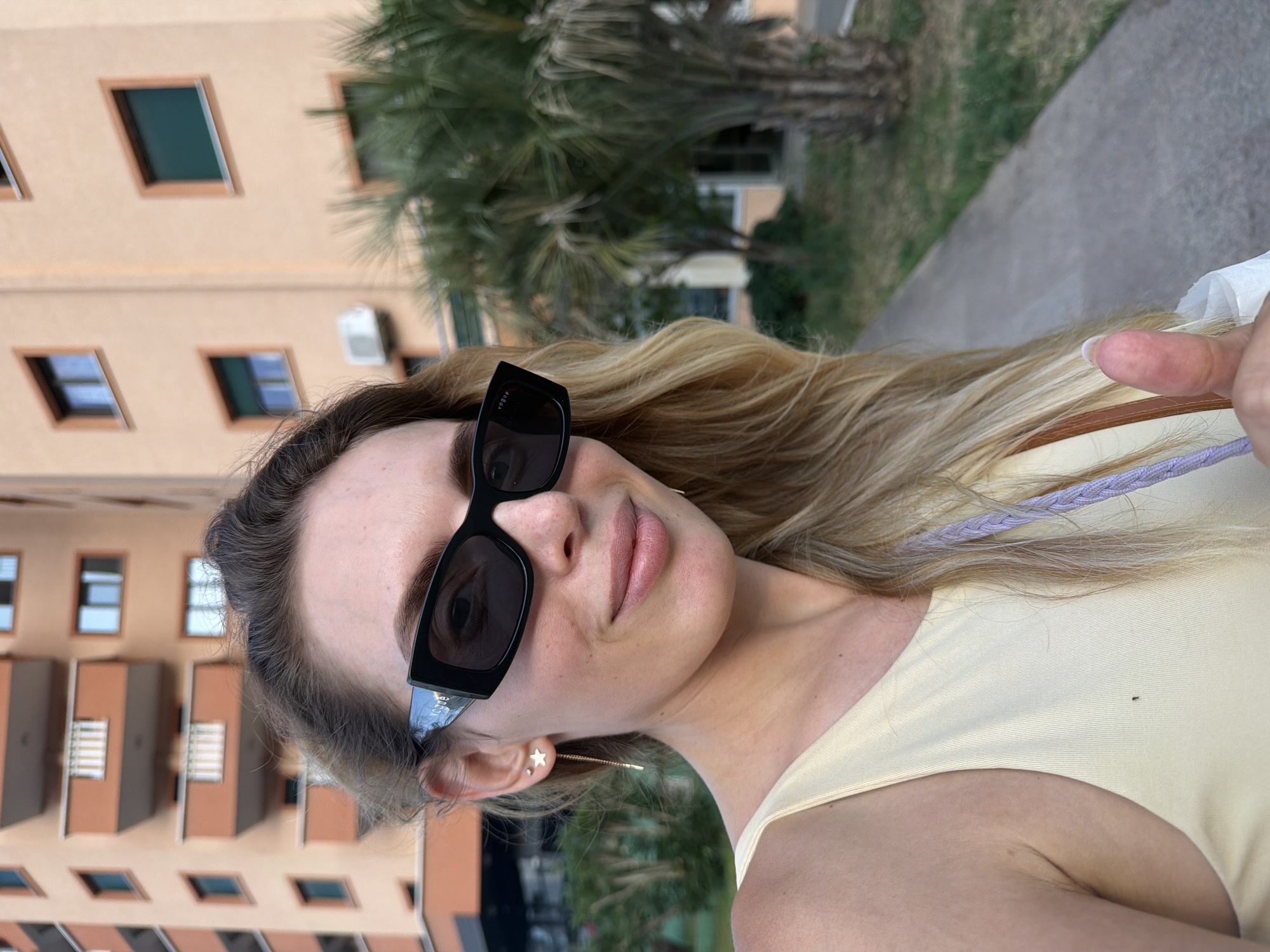 A close-up, waist-up, outdoor selfie taken by a fair-skinned woman with blonde hair and sunglasses, likely in Budva, Montenegro. She is wearing a light yellow tank top and a delicate star-shaped earring with a long chain. Her right hand is visible, giving a thumbs-up gesture. Behind her, a modern apartment building with orange balconies and green-shuttered windows stands against a clear sky. To her right, a palm tree and greenery are visible, suggesting a Mediterranean climate. The woman appears to be walking on a paved path.
