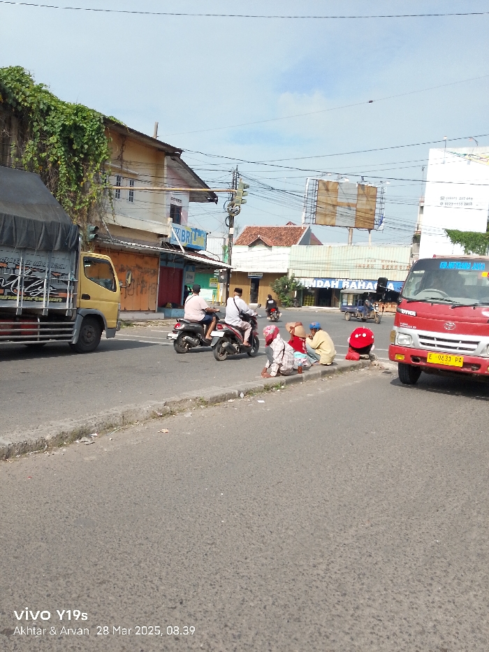 The scene is a street intersection in Jatibarang, Indonesia, at coordinates -6.475, 108.304. A yellow truck is parked on the left side of the road, facing the right. Two motorcycles are parked on the right side of the road. One man is riding a motorcycle, another is standing nearby, and a third is walking in the street. On the right side of the intersection, there are four people sitting on the sidewalk. All of them are wearing traditional clothing. There are various buildings on both sides of the street, with some of the buildings having signs in the Indonesian language. There is a billboard on the right side of the road advertising a "Toko Mitra". The sky is clear with a few wispy clouds. The photo was taken with a VIVO Y19s on March 28, 2025, at 8:39 AM.