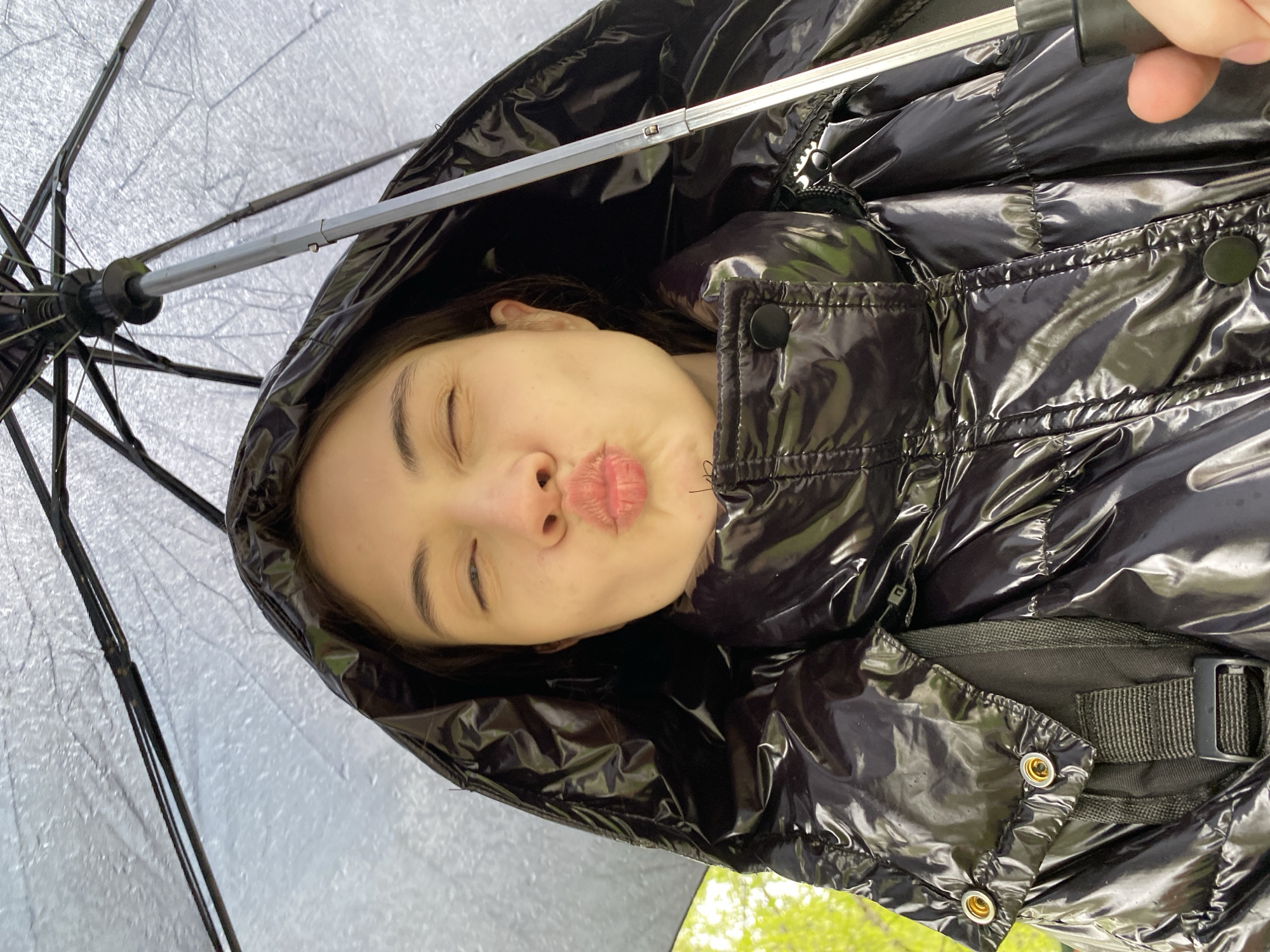 A young woman with fair skin is centered in the frame, tilting her head to the right with her eyes closed and lips pursed in a kissing expression. She is wearing a shiny black puffer jacket with a hood pulled up over her head. An open umbrella is above her, its ribs and translucent canopy visible. The background shows a hint of green foliage. There is no evidence to suggest the location is Votkinsk, Russia.