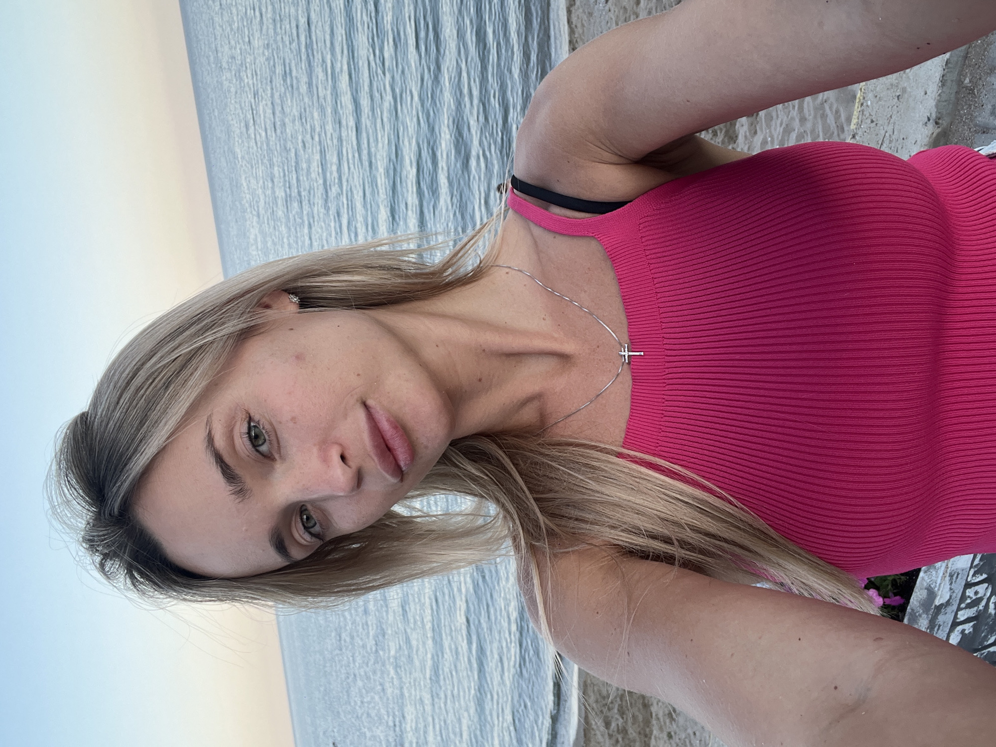 In the city of Fontanka, Ukraine, a young woman with blonde hair and green eyes is taking a selfie on a beach. She is wearing a bright pink ribbed tank top and a silver necklace with a cross pendant. The sea is visible in the background, with gentle waves and a clear sky. The sand is light brown, and there are some rocks and debris on the ground. The woman is looking directly at the camera, with her lips slightly parted. Her hair is pulled back loosely, with strands falling around her face. The overall mood of the image is relaxed and natural, capturing a moment of personal reflection by the sea.