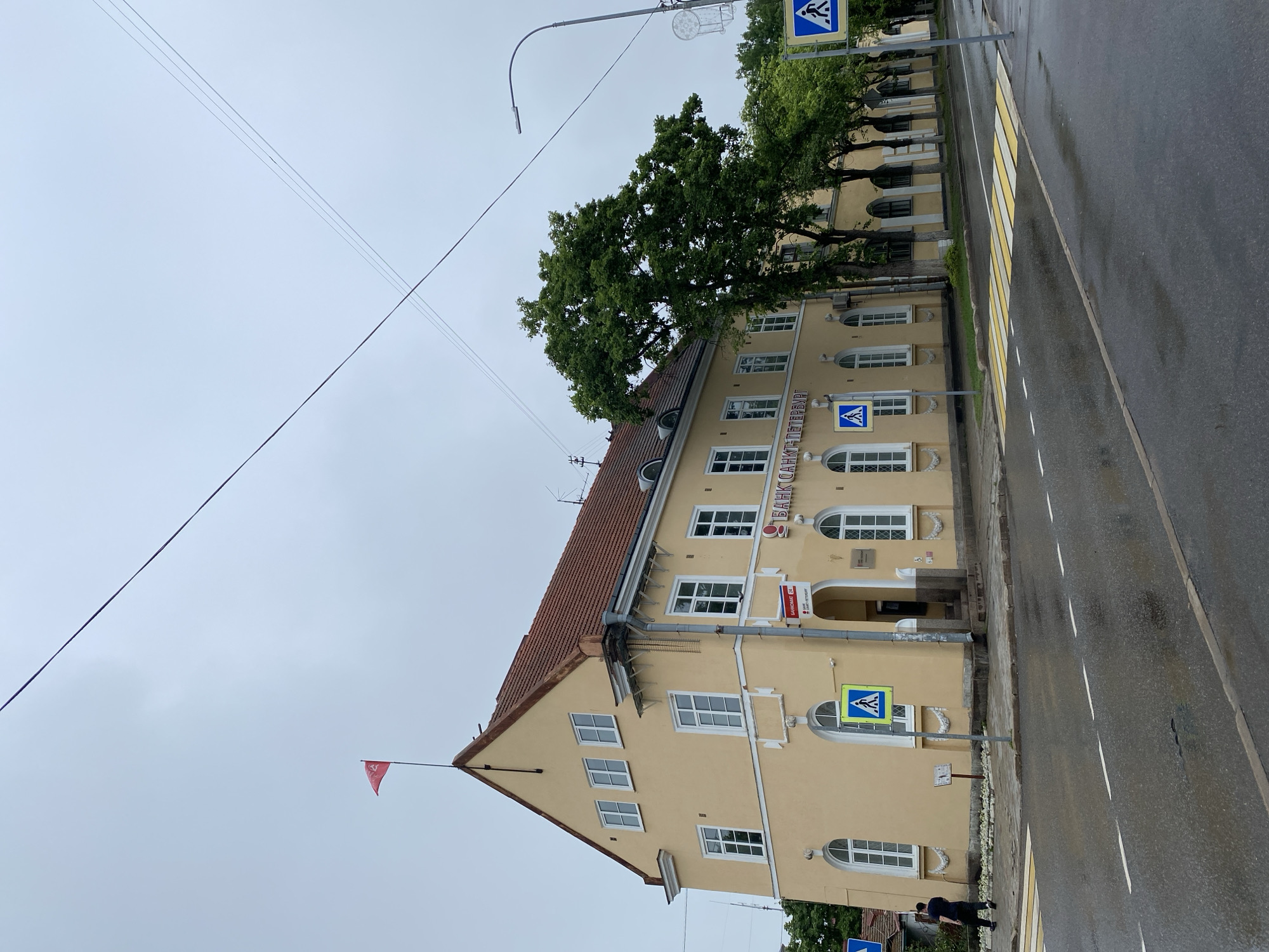 The image shows a yellow building with a red-tiled roof on a cloudy day in the city of Priozersk, Russia. The building has a banner with the words "БАНК САНКТ-ПЕТЕРБУРГ" (Bank Saint Petersburg) written on it. There are several white-framed windows on the building, some of which are arched. A red flag is flying from the rooftop. In front of the building, there is a crosswalk with yellow and white stripes on a wet asphalt road. Two pedestrian crossing signs with blue backgrounds and white figures are visible. A street lamp is also present. A person in a dark shirt and pants is walking away from the camera on the sidewalk. Trees line the street in front of the building. The overall scene depicts a street in a Russian city on a day with overcast skies and recent rain.