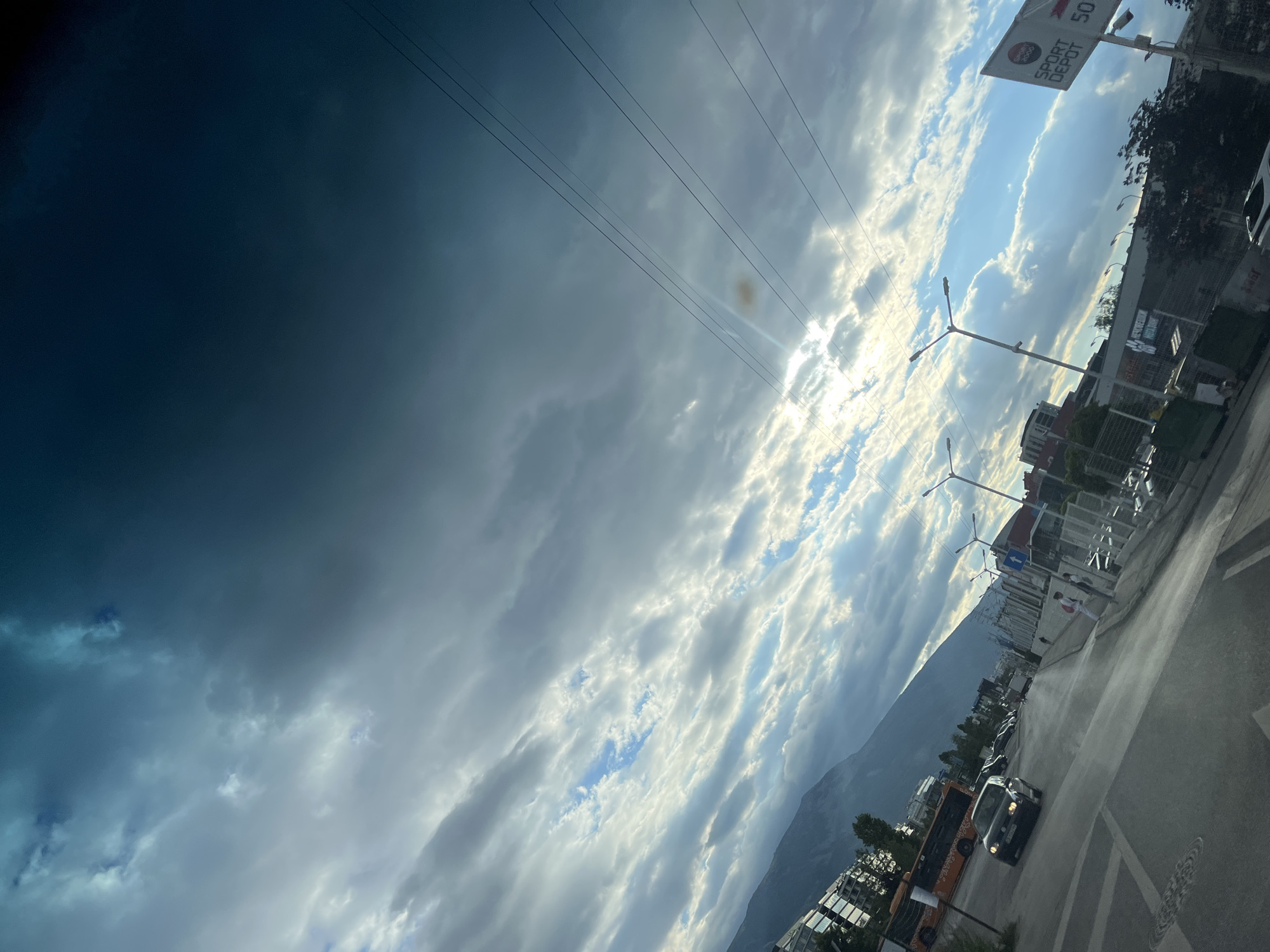 The image shows a street scene with traffic, buildings, and a mountain in the background, under a dramatic sky. A black car is driving on the asphalt road, with other vehicles parked and moving further down. On the left side of the road, an orange bus is visible, along with multi-story buildings and trees. In the background, a large mountain range is silhouetted against the sky. The sky itself is filled with a mix of dark and light clouds, with the sun peeking through, creating streaks of light. There are utility poles and wires crisscrossing the sky. A sign for "SPORT DEPOT 50" is visible on the right side of the image. The overall impression is of a city located near a mountainous region, with active urban life beneath a dynamic sky.