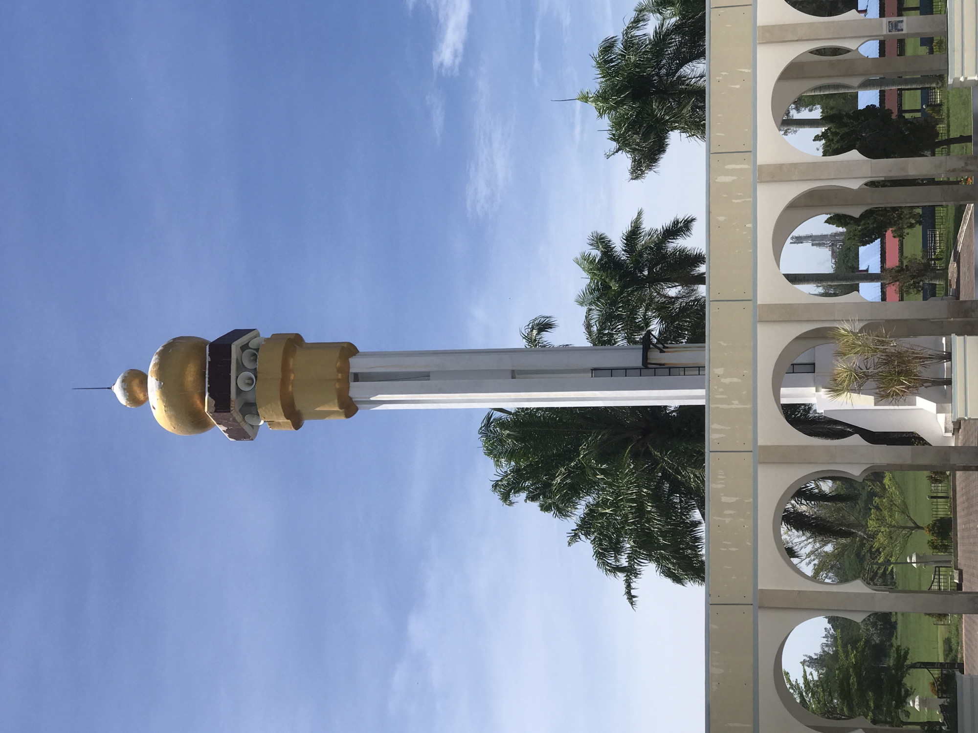 The image depicts a minaret with a golden dome, likely part of a mosque, against a blue sky. The minaret is primarily white, with golden accents. Adjacent to the minaret are palm trees. A white, colonnaded structure with arched openings is visible on the right. The location is Tambontunong, Indonesia.