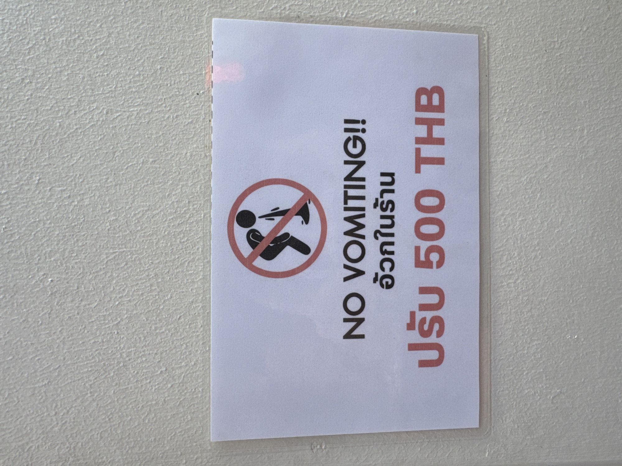 The image shows a sign posted on a white wall. The sign has a "no vomiting" symbol (a person vomiting inside a red circle with a line through it). Next to the symbol, the text "NO VOMITING!!" is printed in black, followed by Thai script. The sign concludes with "ปรับ 500 THB" (Fine 500 THB), indicating a fine of 500 Thai Baht for vomiting. The sign is inside a transparent plastic sleeve or laminate, possibly to protect it. Bangkok, Thailand is not visible in the image.