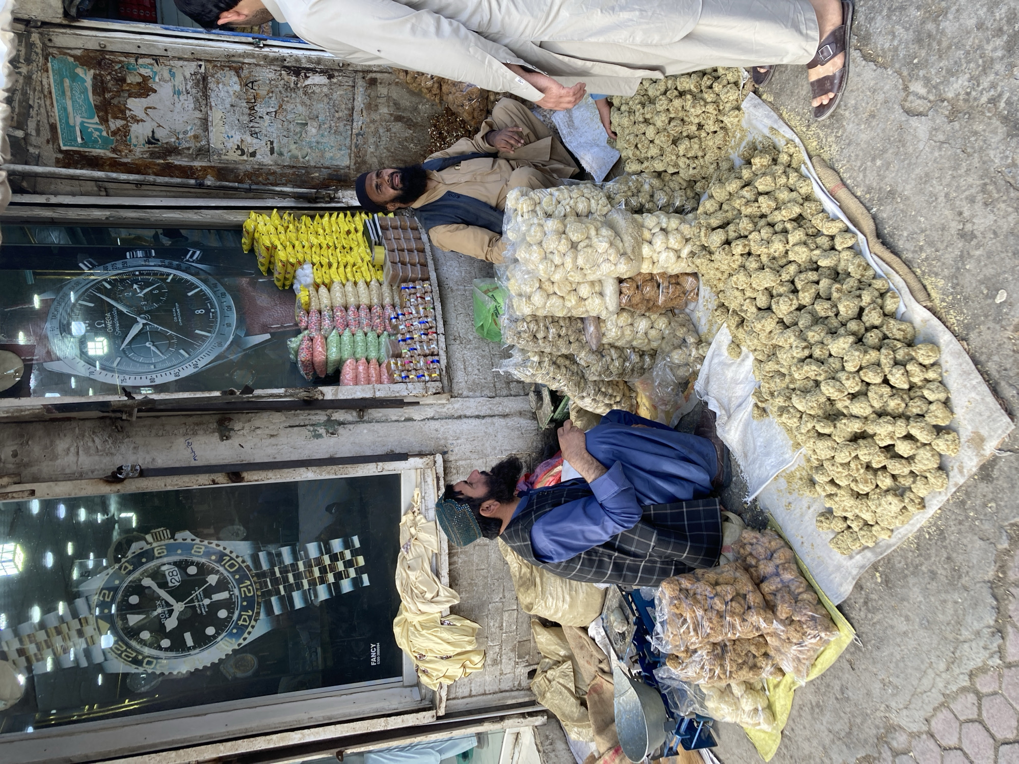 The image shows an outdoor market scene in Quetta City Tehsil, Pakistan. Two men with beards are reclining on the ground. One is dressed in a light brown shalwar kameez with a dark vest, while the other wears a blue shalwar kameez with a dark checkered vest and a patterned cap. The men are surrounded by piles of what appears to be various types of sweets or snacks, some packaged in plastic and others loose. Above the men, there are storefronts with window displays featuring large posters of wristwatches. One poster is of an Omega Speedmaster watch, and the other is of a Rolex GMT-Master II. The storefront facade is old and shows signs of wear. A person's legs, clad in light-colored clothing, and sandals are visible at the top of the frame. The ground is a mix of concrete and debris.