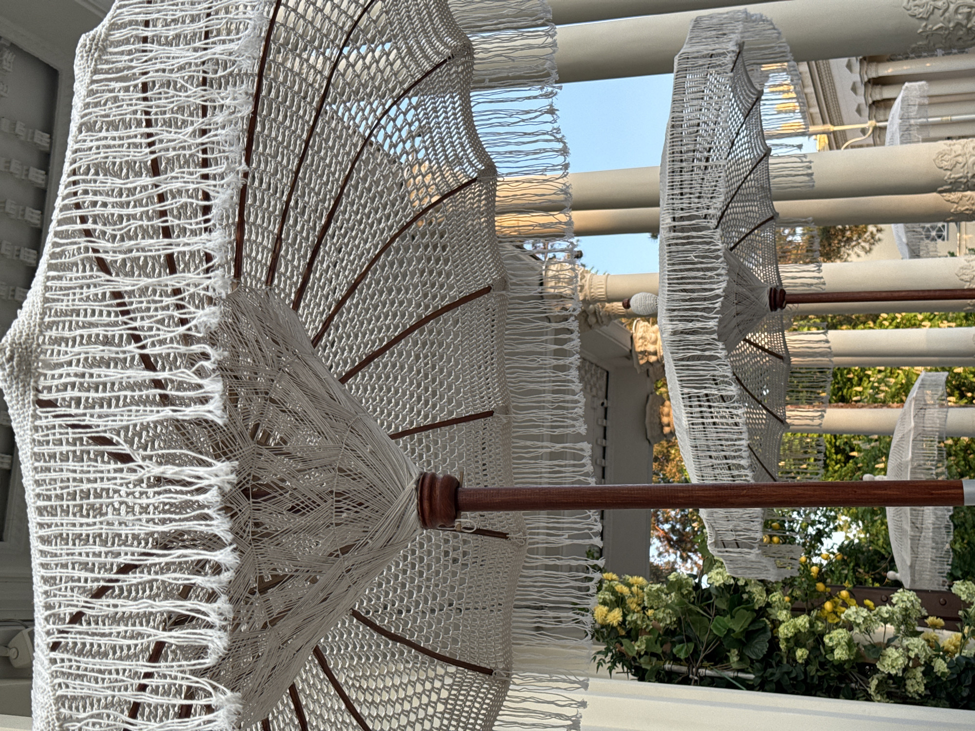 The image shows a row of ornate, white-laced parasols with wooden handles, likely decorations, hanging from a white structure. The background includes blue sky and glimpses of greenery. The parasols are the primary focus, and their intricate design is visible. The setting seems to be an outdoor space, potentially a patio or garden area. It is impossible to determine the location or if it is in Moscow, Russia, based on the image provided.