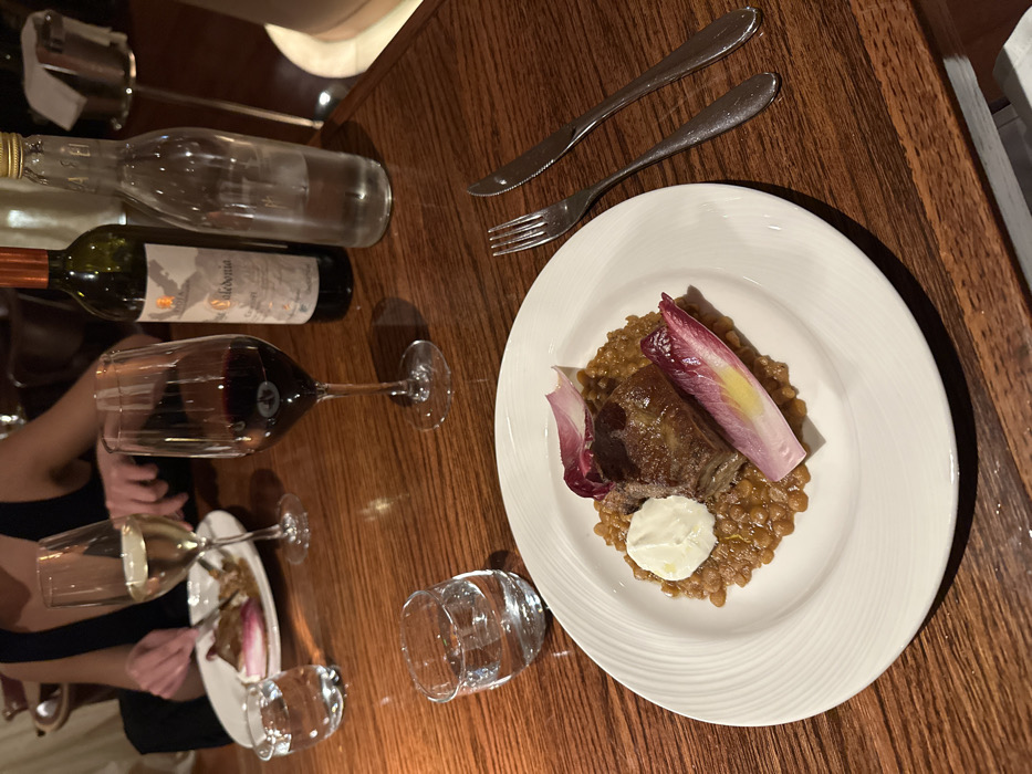 The image depicts an indoor dining scene at a high-end restaurant in the city of London, United Kingdom, featuring a polished wooden table under dim, warm ambient lighting. In the foreground, a white ceramic plate with a fluted rim holds a main course: a rectangular portion of dark, braised meat atop a bed of brown lentils, garnished with a dollop of white sauce and two red and white radicchio leaves. To the right of this plate, a stainless steel fork and knife are positioned. A small, clear glass tumbler containing water sits to the left of the plate. In the mid-ground, a tall-stemmed wine glass holds red wine, approximately one-third full. Adjacent to it, a dark green glass bottle with a cork stopper is visible, partially obscured but showing "Caledonia" on its label. Next to the wine bottle stands a clear glass bottle with a metal screw cap, containing water. In the background, a second individual is partially visible, wearing a dark garment, with hands holding a fork and knife over a white plate containing food. A tall-stemmed wine glass with white wine, approximately one-third full, and another small clear glass tumbler with water are present near this individual.