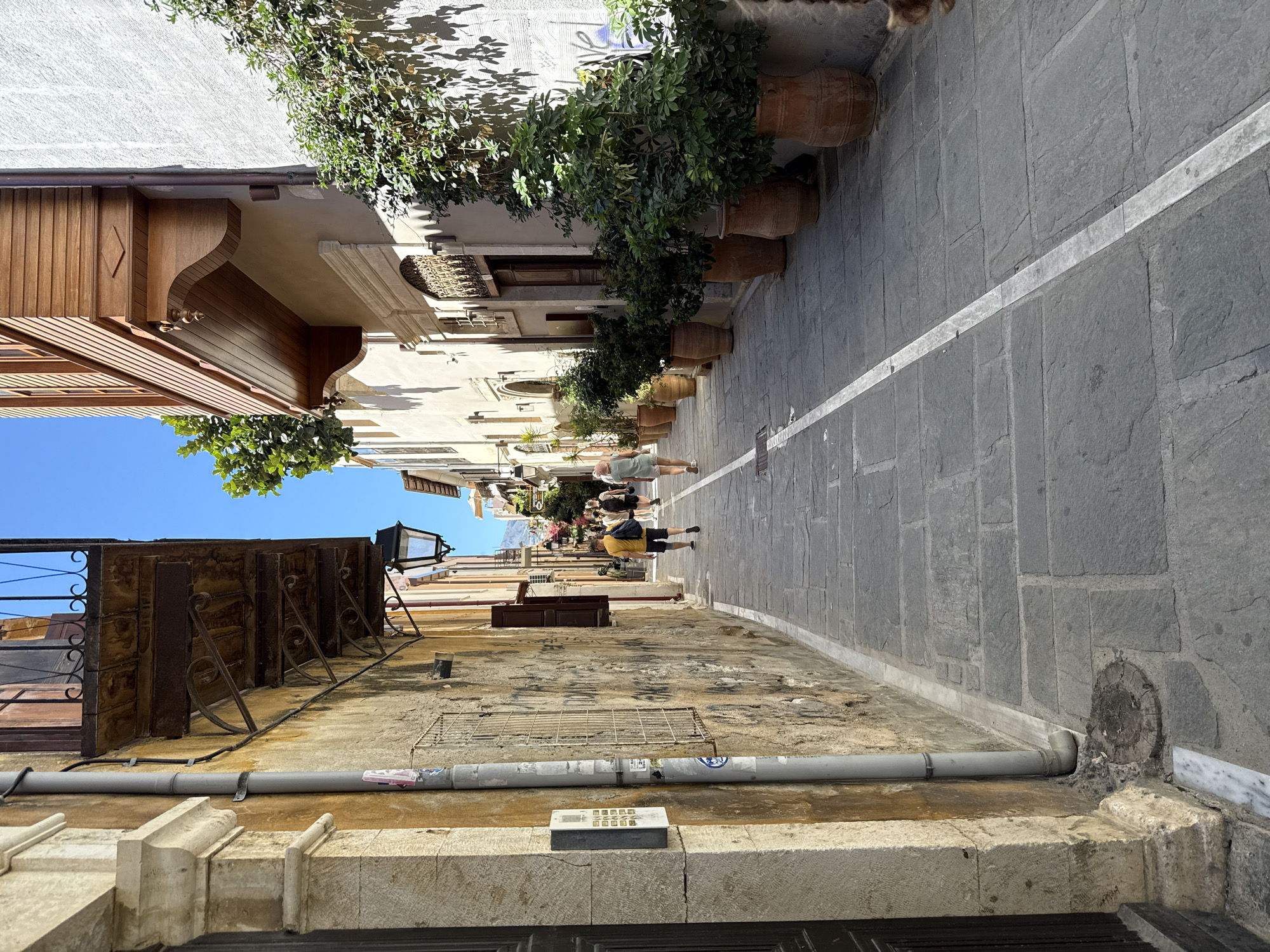 The image depicts a narrow, stone-paved alleyway in Rethymno, Greece, bathed in sunlight. The buildings flanking the alley are constructed of weathered stone and plaster, with wooden balconies and ornate architectural details. The left side of the alley features a rough-textured stone wall with a downspout and an intercom panel. The right side of the alley is lined with large terracotta pots containing lush green plants. Several people are walking away from the viewer down the alley, dressed in casual summer clothing. The ground is paved with large, irregular stone slabs with a white line painted down the center of the alley. The sky is clear blue.