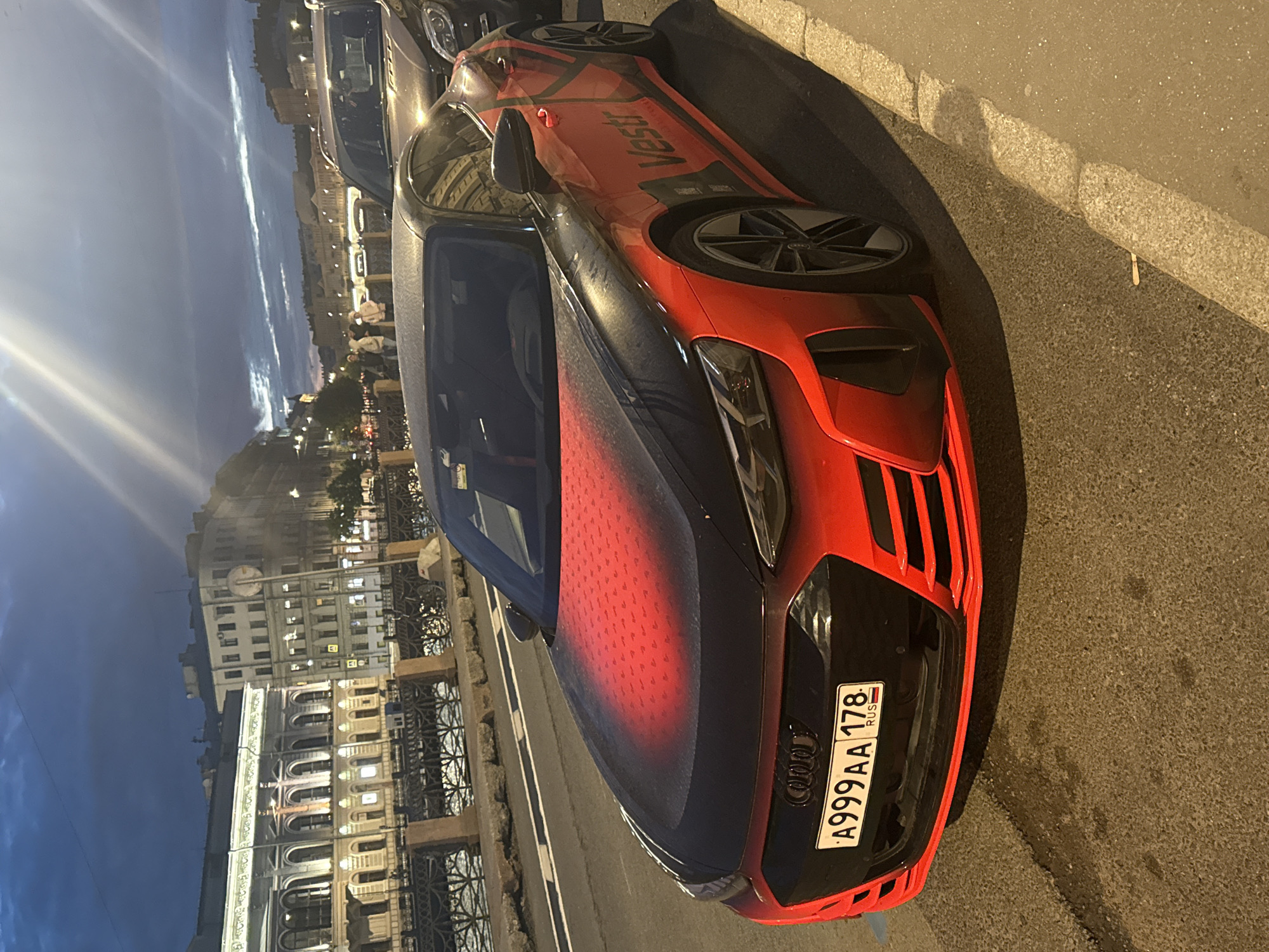 A black and red Audi e-tron GT electric sports car is parked on the right side of the road, facing left. The car has a custom vinyl wrap with a gradient of red on the hood and a logo on the side that reads "Vestr." The license plate reads "A999AA 178 RUS." In the background, there are ornate buildings along a canal, characteristic of St. Petersburg, Russia. The sky is dark with clouds, indicating it is either early morning or evening. There are a few people walking on the sidewalk in the distance, and another car is parked behind the Audi. The scene appears to be on a street with a sidewalk and a stone embankment lining the canal.