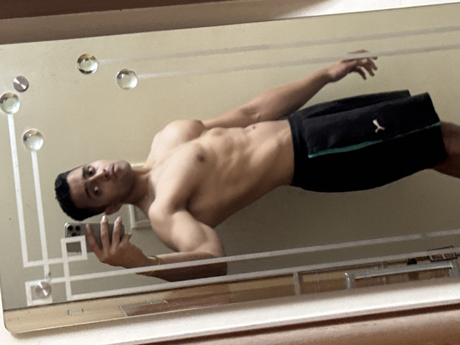 A shirtless man is reflected in a mirror, taking a selfie. He is wearing black shorts with a white Puma logo. His muscular physique is visible, including his abs, biceps, and chest. The mirror has decorative embellishments, including round glass or crystal accents and a geometric pattern near the bottom. The background is a plain, light-colored wall.