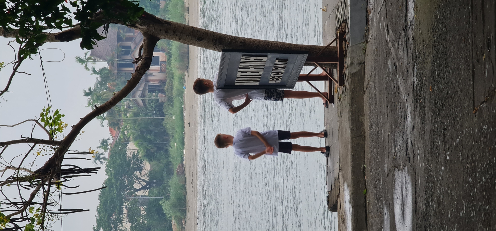 Two young boys stand on a paved embankment near a body of water, posing beneath a sign that reads "DAI TIEN RIVER RESTAURANT." The scene is likely in Hội An Ward, Vietnam. The embankment is adjacent to a beach, visible across the water. A tree branch extends into the frame from the left side, partially obscuring a view of buildings and lush foliage in the background. The sky is overcast.