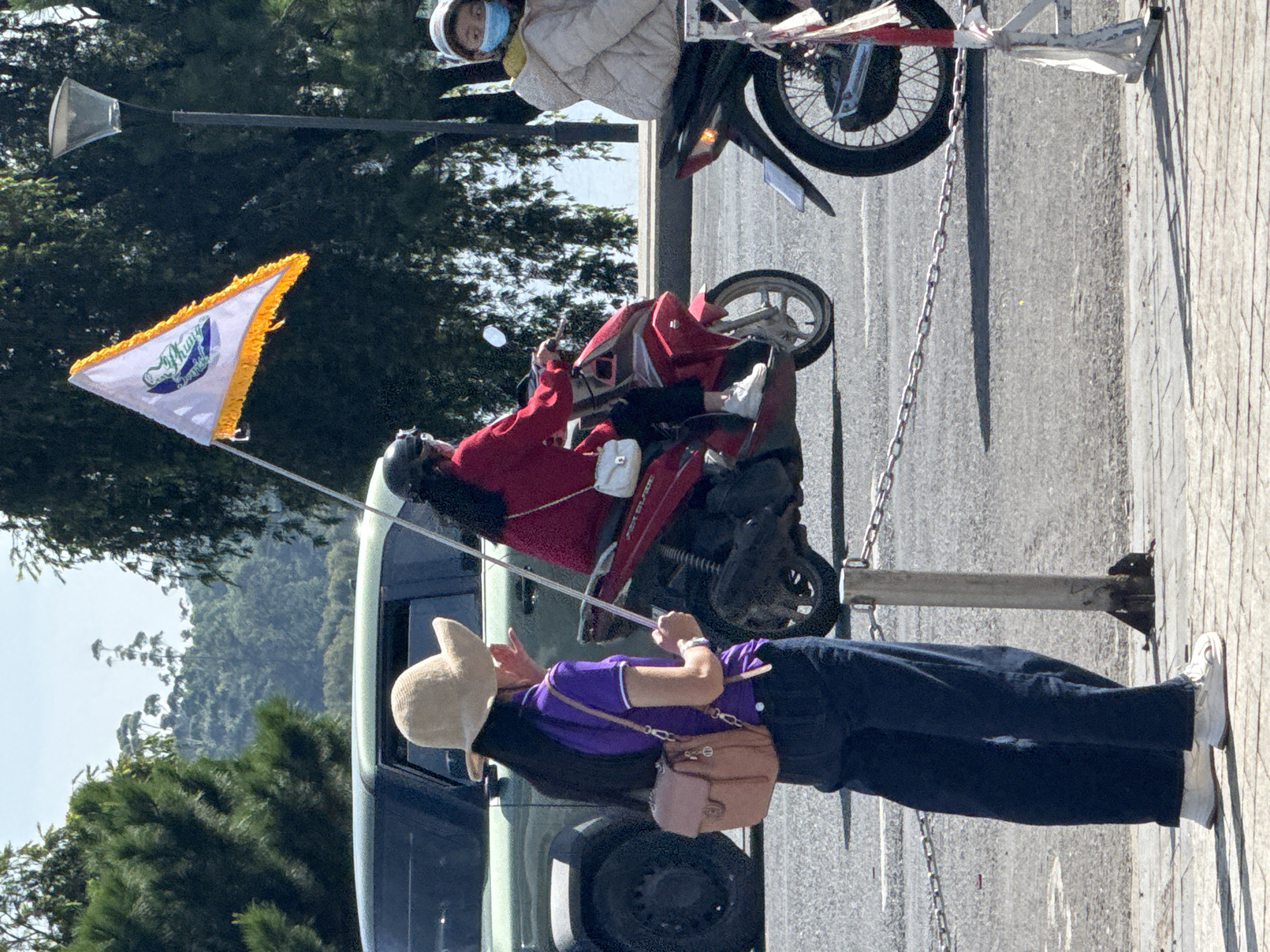 In Da Lat, Vietnam, a woman in a beige hat and purple shirt stands on a tiled walkway, holding a white flag with a yellow fringe. A light brown crossbody bag is slung over her shoulder. Behind her, a person on a red scooter, wearing a red jacket and helmet, holds the flag. To the right, another person on a motorcycle, wearing a light-colored jacket and a blue face mask, is partially visible. A dark green SUV is parked behind the woman in the foreground. The scene appears to be near a street with a body of water in the background, possibly Xuan Huong Lake.