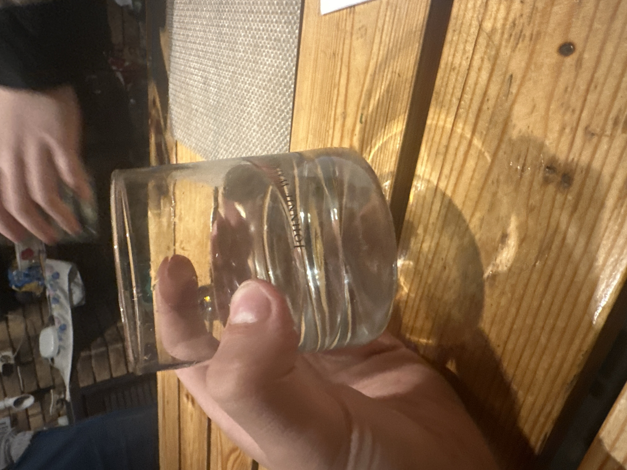 A hand holds a clear glass of clear liquid, possibly water or a light-colored alcoholic beverage, on a wooden table. The glass has a ribbed texture and a logo or text that reads "JOHNNIE WAL..." is visible on the side of the glass. In the background, another person's hands are visible, also interacting with glassware. The environment appears to be an indoor setting with a brick wall and a patterned placemat on the table. The image is taken from a slightly low angle, focusing on the glass and the hand holding it. The lighting suggests an indoor evening setting. This scene is likely taking place in a private residence or a casual dining establishment in Востец Снт, Russia.