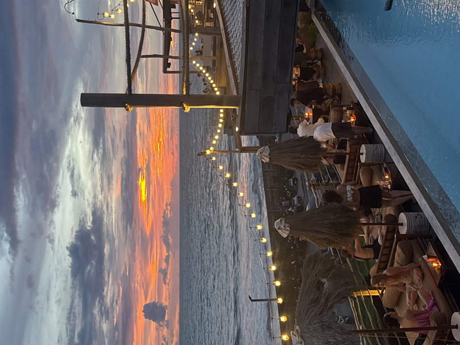 This image captures an elevated outdoor lounge and dining establishment situated on the coast in Canggu, Indonesia, during sunset. The sky displays a dramatic sunset with a vibrant orange and red horizon, transitioning into darker grey and purple clouds overhead. The ocean is dark blue-grey, exhibiting visible waves breaking near the shore.
In the foreground, a clear blue infinity pool occupies the lower right quadrant. Adjacent to it and extending to the left, a multi-level outdoor seating area is visible. The lower tier contains several lounge settings comprising cushioned daybeds and small tables. Three large, conical straw-thatched umbrellas, two topped with white covers, provide shade. Approximately ten to fifteen individuals, dressed in casual attire, are present across these areas, engaged in sitting, lounging, and social interaction. Specific groups are visible on daybeds and around tables.
The establishment features dark wooden architectural elements, including a tall vertical post and horizontal beams with rope detailing on the right side, suggesting a nautical theme. Multiple strings of warm-toned, bulbous light bulbs illuminate the structures and extend along the ocean-facing railing. Below the main lounge area, a narrow coastal road runs parallel to the shore, where several motorbikes are parked. Small, glowing lanterns or candles are placed on the tables within the lounge areas.