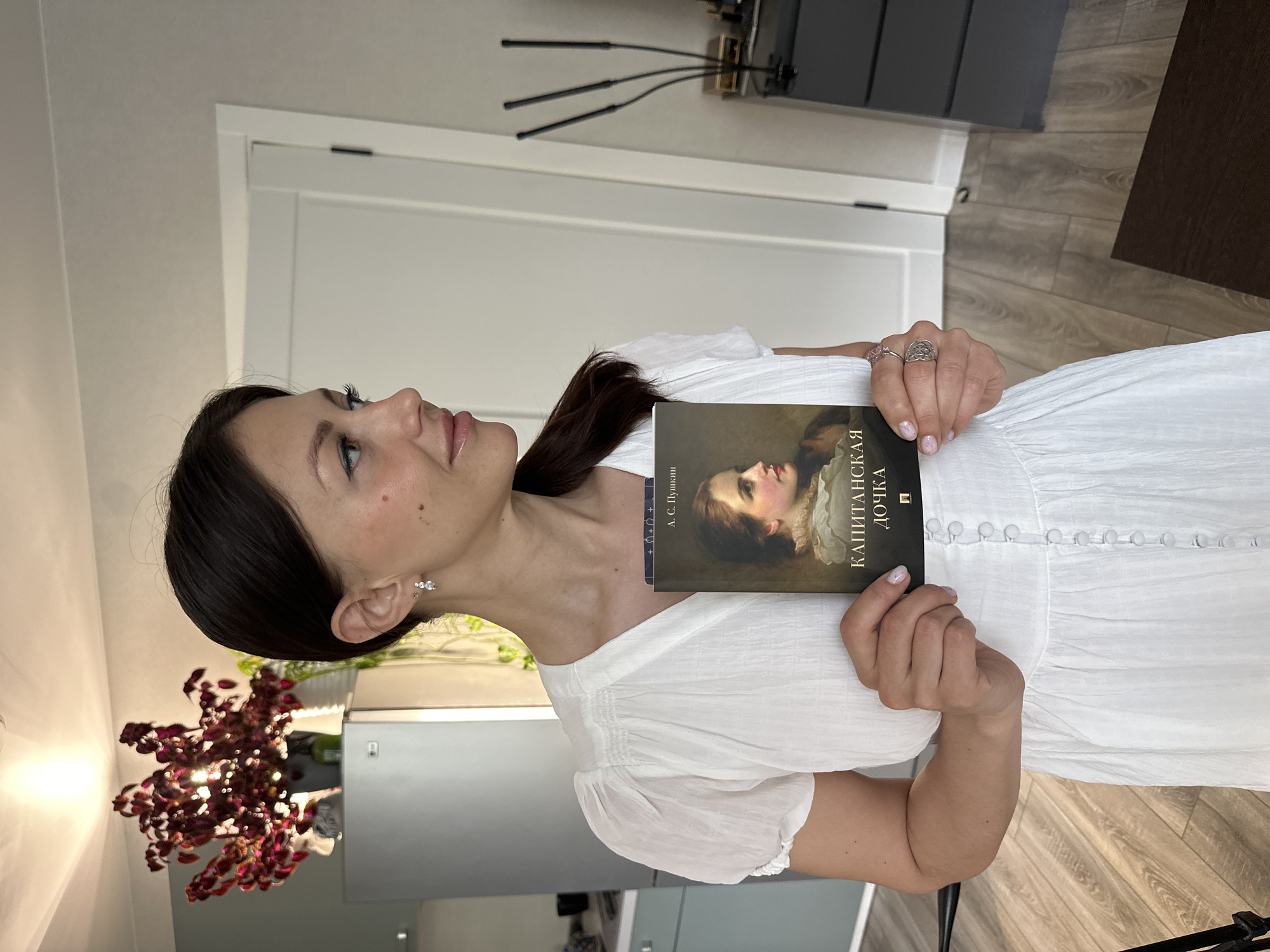 A woman in a white dress is holding a book titled "Капитанская дочка" (The Captain's Daughter) by A. S. Pushkin. She is indoors, likely in an apartment or home. The background includes a door, some furniture, and a lighting fixture. There are also flowers visible. The location is likely Murino, Russia, given the book is in Russian.