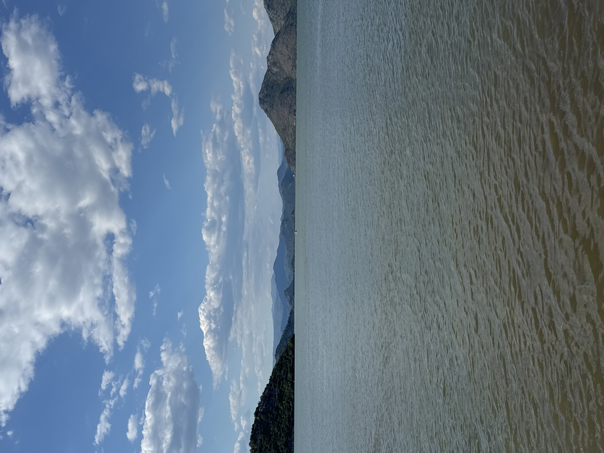 The image depicts a serene landscape dominated by a large body of water. In the foreground, the water exhibits gentle ripples, suggesting a calm atmosphere. The middle ground is occupied by the vast expanse of the lake, reflecting the sky and surrounding mountains. To the left, a lush green, tree-covered hill slopes down towards the water, its form softened by distance. Further in the distance, a series of undulating mountains form the backdrop, their rugged textures visible against the clear blue sky. The sky itself is a vibrant blue, dotted with numerous white, fluffy clouds. A small boat is visible on the water in the far distance, adding a sense of scale to the expansive scenery. The overall impression is one of natural beauty and tranquility. The scene is located in Seoca, Montenegro.