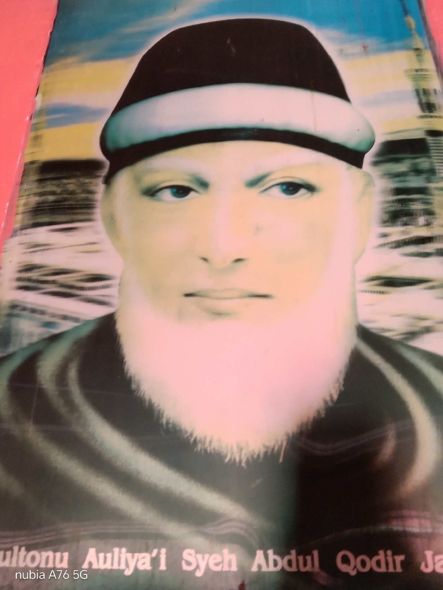 The image is a close-up, front-facing portrait of a man wearing a black and white kufi hat. He has fair skin, blue eyes, and a prominent white beard. The background is a blurred abstract pattern with hints of blue and yellow. Below the portrait, text is visible, reading "ultonu Auliya'i Syeh Abdul Qodir Ja" and "nubia A76 5G". The overall image quality is poor, with low resolution and oversaturation, giving it a vintage or faded appearance. The context suggests this might be a photograph displayed in a public or personal space in Cakung, Indonesia, possibly a poster or artwork. There are no other individuals or specific environmental details of Cakung depicted in the image.