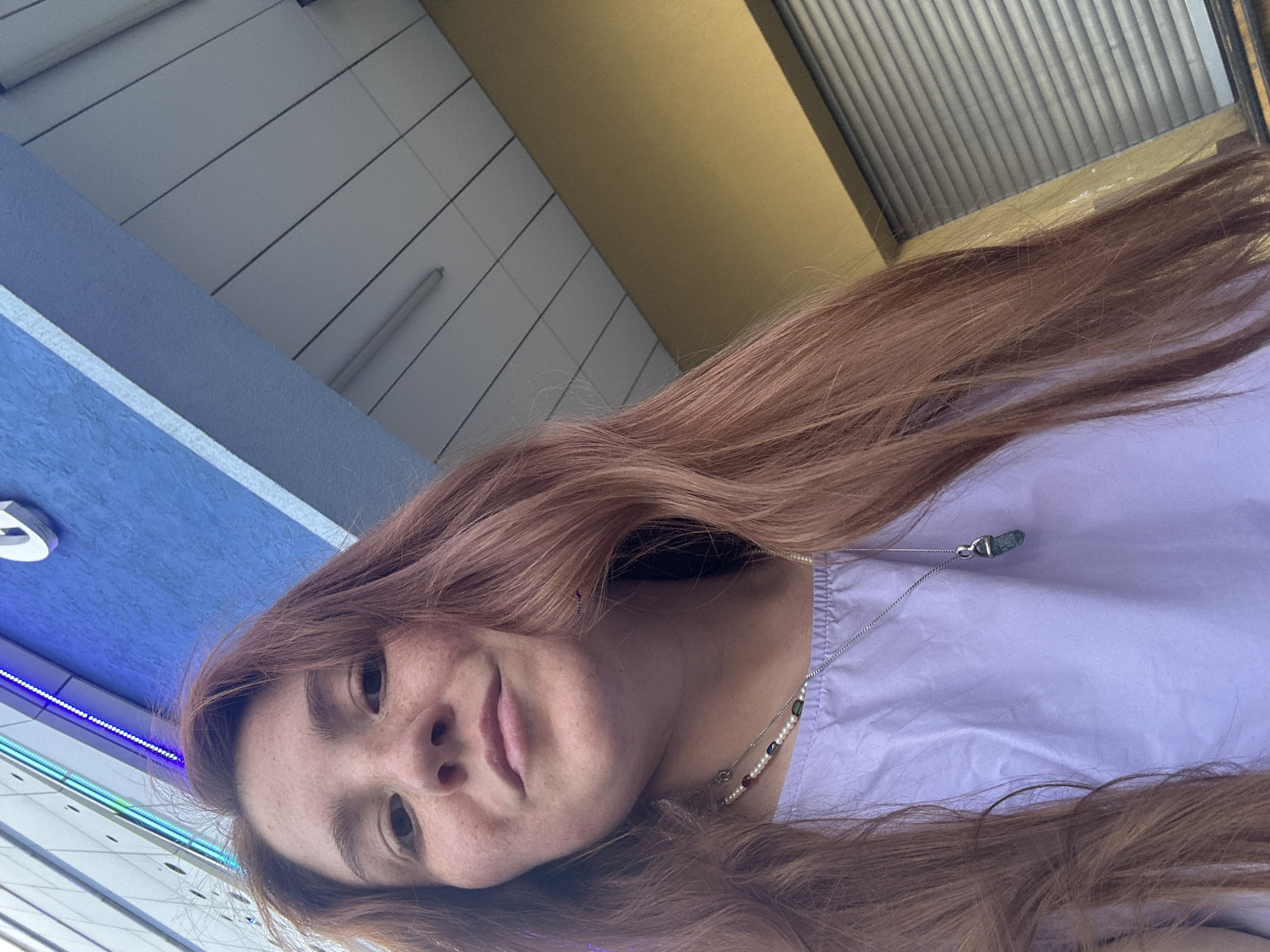 A young woman with auburn hair and freckles is shown in a selfie from a low angle. She is wearing a light purple top and a beaded necklace with a pendant. Behind her, a blue building with a colorful light strip is visible, along with a section of a yellow building and a gray rolled-down shutter. The background suggests an urban environment, possibly a street or a commercial area. The image does not provide any specific visual cues to indicate a location in Nizhny Novgorod, Russia.