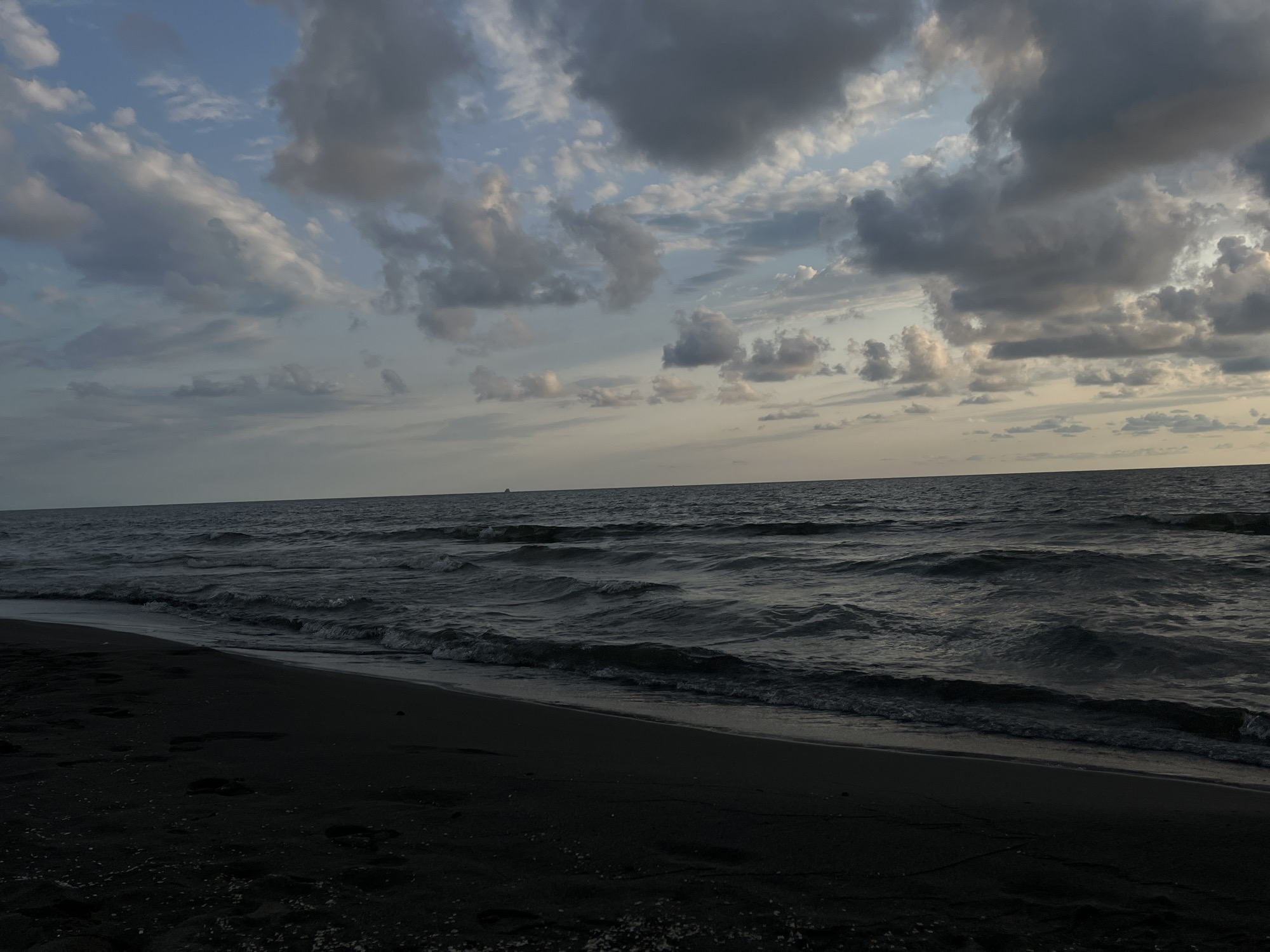 The image depicts a dark, sandy beach bordering a dark, choppy sea under a cloudy sky. The foreground is dominated by the black sand, with some scattered lighter debris or shells. Footprints are visible in the sand near the water's edge. The sea is a deep, muted blue-gray, with whitecaps and ripples indicating moderate wave action. In the distance, a faint, dark silhouette of a vessel is visible on the horizon. The sky is a mix of pale blues and grays, with large, textured clouds that suggest a late afternoon or early evening setting. The overall lighting is dim, creating a somber and atmospheric mood. This scene is likely located on the Black Sea coast of Georgia, possibly in the vicinity of სუფსის თემი.