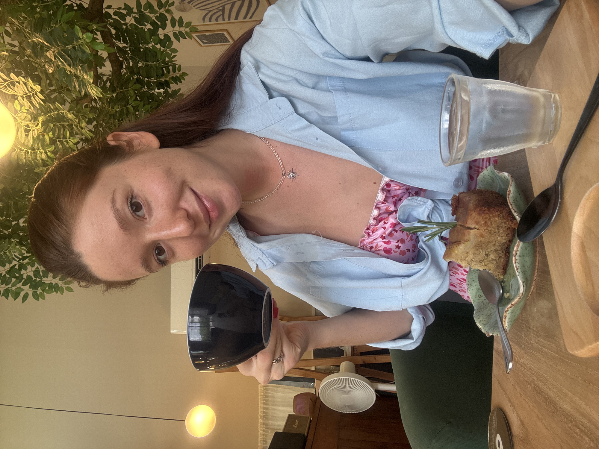 The image, oriented vertically, captures a woman with long brown hair and a gentle smile, seated at a wooden table in an indoor cafe or restaurant setting. She is looking towards the viewer, holding a black ceramic mug in her right hand. She wears a light blue button-up shirt unbuttoned over a pink top with a floral pattern, and dark green pants. A silver necklace with a star-shaped pendant adorns her neck, and a ring is visible on her left ring finger.
On the table in front of her, a clear glass filled with a beverage, possibly iced, is placed alongside a rustic slice of cake or sweet bread. The cake, garnished with a sprig of rosemary, rests on a unique green, organically-shaped plate. A silver spoon and a black serving spoon are also on the table.
The background features decorative elements, including lush green artificial plants, creating a natural and inviting atmosphere. Soft, warm light emanates from spherical pendant lamps, suggesting evening or late afternoon ambiance. A white standing fan is visible further back, hinting at a warm climate, which aligns with the provided location context of Choeng Thale, Thailand. Other dark furniture pieces, including what appears to be a wooden cabinet, are subtly present in the background. The scene conveys a relaxed and enjoyable moment. No visible text is present in the image.