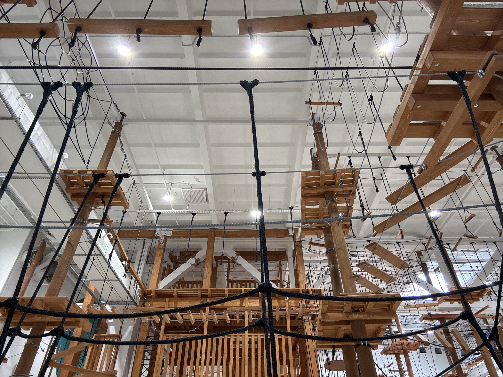 This is an image of an indoor ropes course and adventure park. The structure is made of wooden platforms, beams, and numerous ropes and cables of various thicknesses. The course appears to be multi-level, with different challenges and obstacles suspended at varying heights. The ceiling is white and industrial, with visible light fixtures and ventilation systems. The image is taken from a low angle, looking upwards into the intricate network of the ropes course. There are no people visible in the image. The ropes course is located at the "Park of the Russian Geographical Society" in Orenburg, Russia.