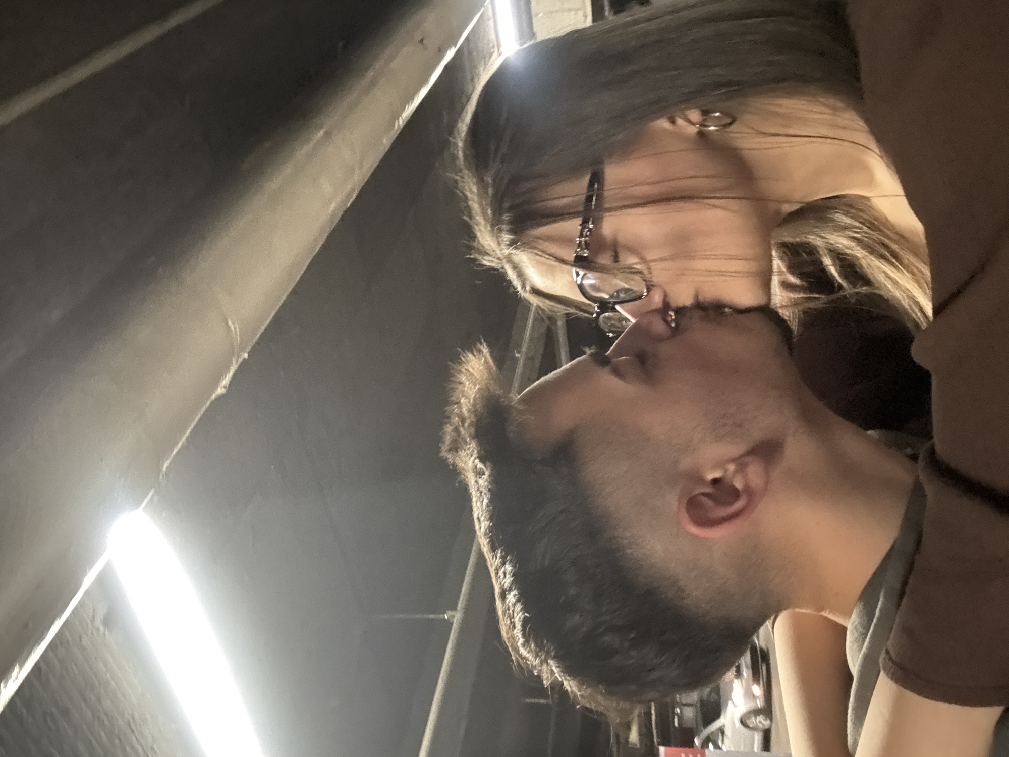 A couple is kissing, with the woman wearing glasses and a nose ring. The location appears to be under an overpass or elevated structure, potentially in Sofia, Bulgaria based on visual cues such as architectural styles or traffic patterns. The lighting is a mix of ambient and artificial, suggesting an urban environment at night or in a dim area.