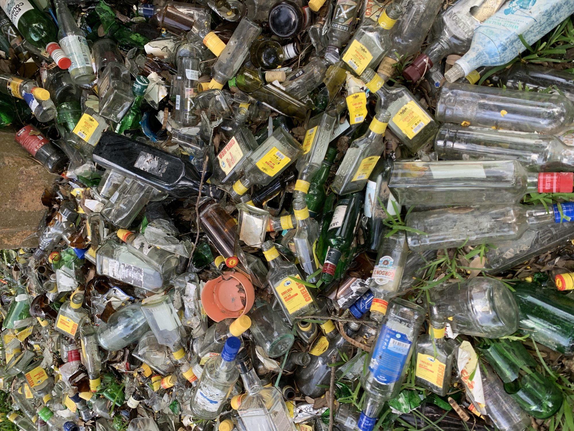 This is a top-down, medium shot of a large pile of discarded glass bottles, primarily alcohol bottles. The bottles are of various shapes, sizes, and colors, including clear, green, and brown glass. Many of the bottles are empty, while some appear to be partially filled. Several bottles have labels that are partially legible, with brands like "CHELSEA," "SMIRNOFF," "GORDON'S," "SQUADRON," and "WHITE WALKER" being identifiable. There are also broken pieces of glass scattered throughout the pile, indicating that some bottles have been smashed. The scene appears to be outdoors, with some dirt, leaves, and blades of grass visible amongst the bottles, suggesting it's a natural or semi-natural setting like a roadside or an abandoned area. The lighting suggests it could be daytime. There are no people visible in the image, and the overall impression is one of neglect and waste. The presence of so many empty bottles suggests a past event or ongoing consumption. The location context is Jos, Nigeria.
