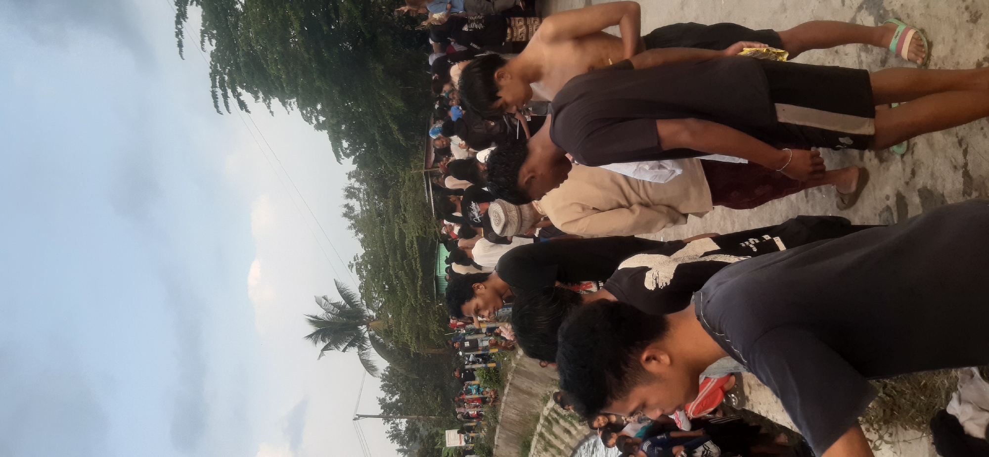 The image depicts a gathering of people outdoors, likely in the city of Sepakek, Indonesia, given the context. The scene appears to be near a body of water, possibly a river or canal, with a concrete embankment visible on the left. A crowd of individuals, predominantly young men and some women and children, are standing around. Some are looking in various directions, while others appear to be engaged in conversation.
The environment features lush green trees and vegetation in the background, suggesting a tropical or subtropical climate. The sky is overcast with some lighter clouds. Various clothing styles are visible, including t-shirts, shorts, and some traditional attire. There are also motorcycles parked in the background, indicating transportation.
The overall atmosphere seems to be that of a community event or gathering, with people milling about on a paved area next to the water. The composition of the image is vertical, capturing a segment of the crowd and the surrounding environment.