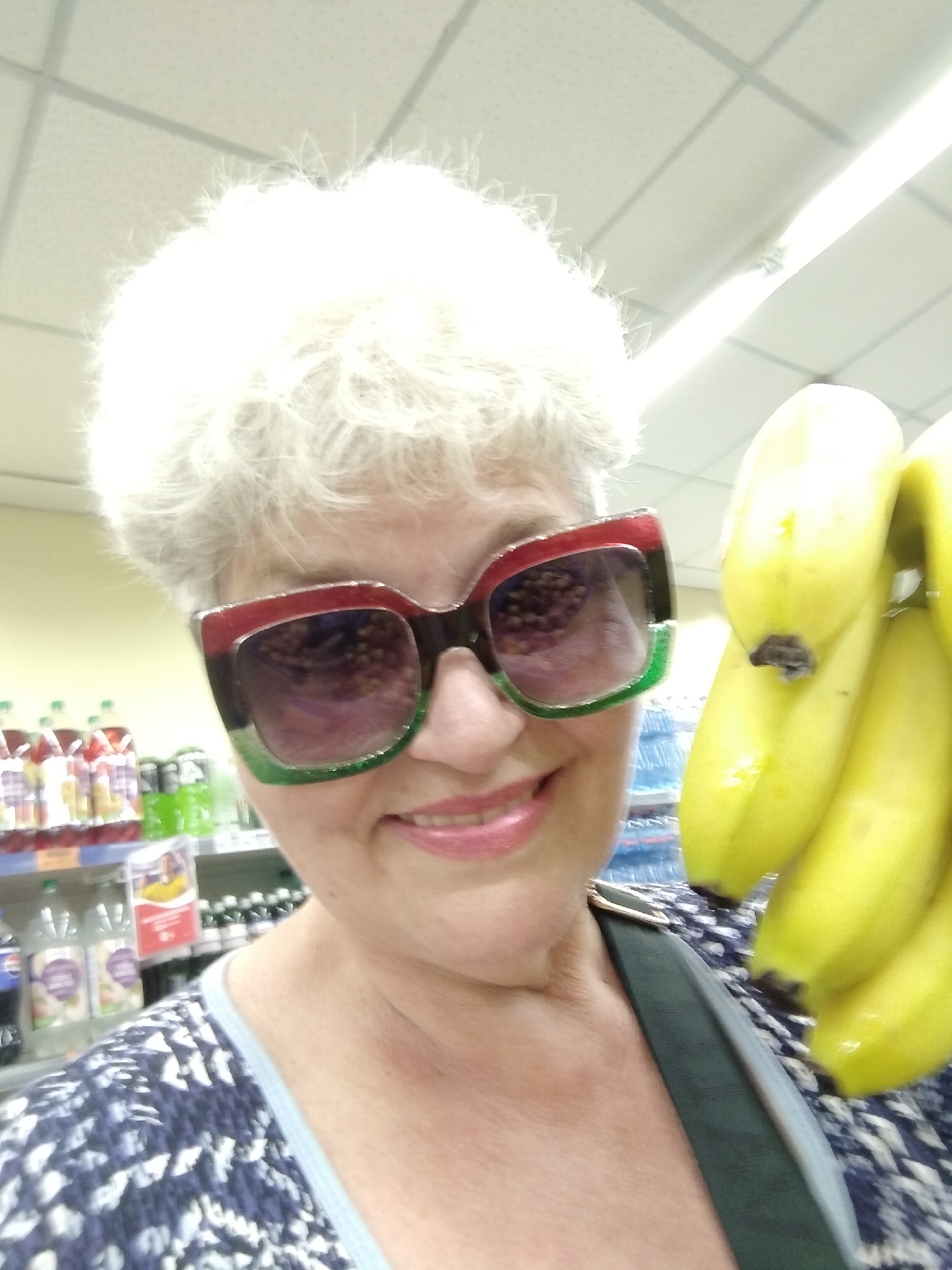 A medium shot captures a woman with short, white, curly hair wearing large, square, multi-colored sunglasses with red and green rims. She is holding a bunch of bright yellow bananas in her right hand, which are positioned in the foreground and partially obscure her face. The woman is smiling and wearing a blue and white patterned top with a light blue neckline. The background appears to be a retail environment, likely a grocery store in Horishni Plavni, Ukraine, with shelves of beverages visible on the left and a ceiling with fluorescent lighting fixtures. The overall impression is that of a candid photograph taken while shopping.