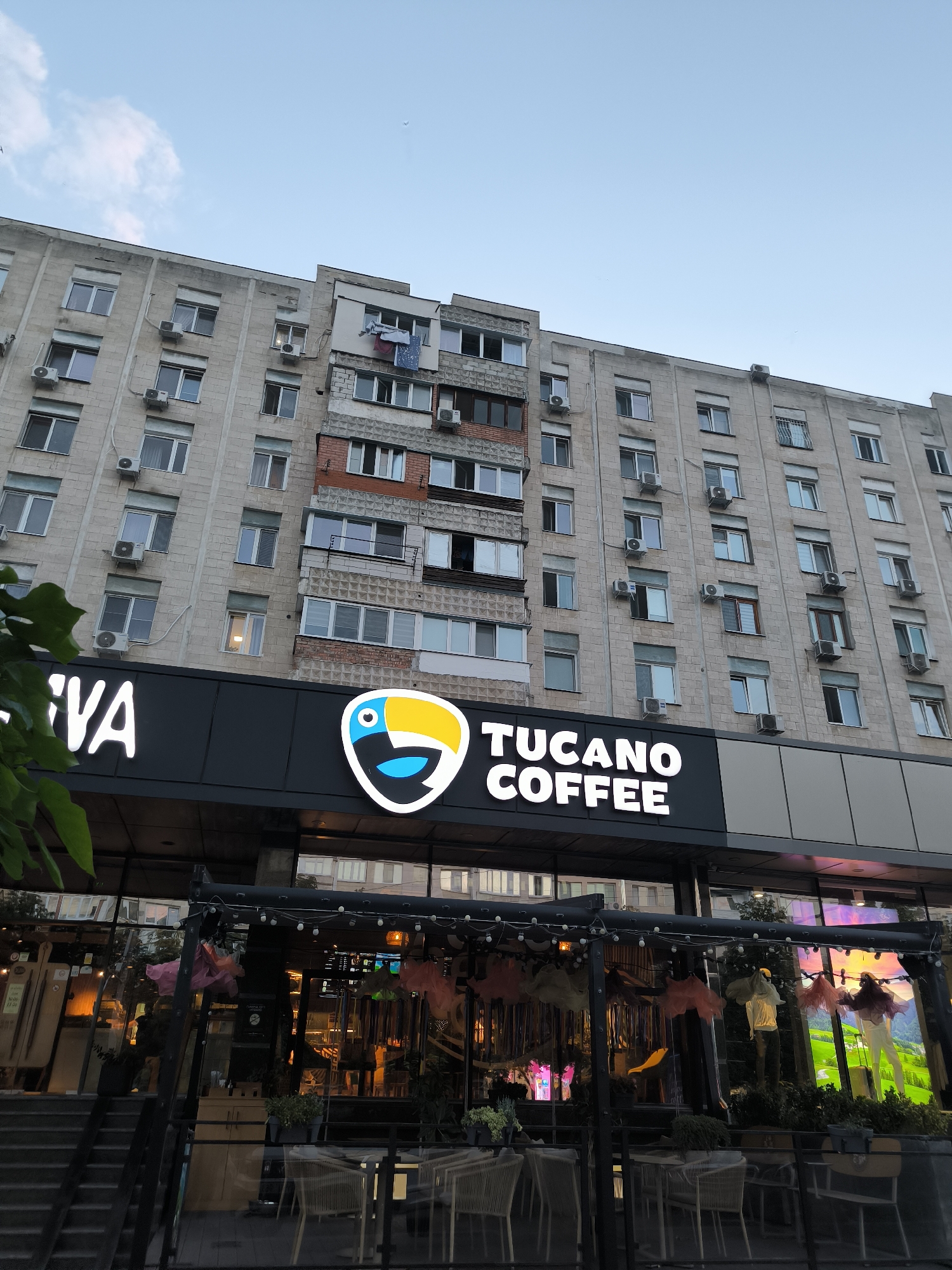 This is a medium-angle, eye-level shot of a building facade with a cafe on the ground floor. The building is a multi-story residential building with a light-colored stone exterior, numerous windows, and air conditioning units visible on each floor. On the ground floor, a cafe with the name "TUCANO COFFEE" is prominently displayed in white lettering against a black background. The cafe has large glass windows and an outdoor seating area with tables and chairs. The overall environment appears to be an urban setting, likely in Chișinău, Moldova, given the architectural style and the presence of a popular coffee chain. The sky is overcast and appears to be early evening. There are no people visible in the scene.