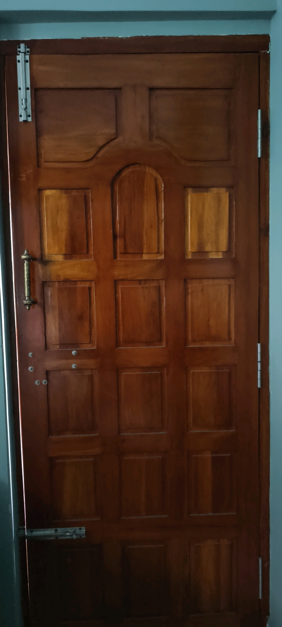 The image shows a closed, wooden interior door with multiple panels. The door is brown and has a traditional design with raised panels and decorative molding at the top. A brass-colored door handle is visible on the left side of the door, along with a silver deadbolt lock. Two silver hinges are visible on the right edge of the door, suggesting it opens inwards. The door is mounted in a frame that appears to be painted a light blue or gray. There is a white bolt latch near the top left of the door. The lighting suggests an indoor environment, possibly a hallway or room. The image does not contain any discernible geographical features that would indicate its specific location within Kundrathur, India.